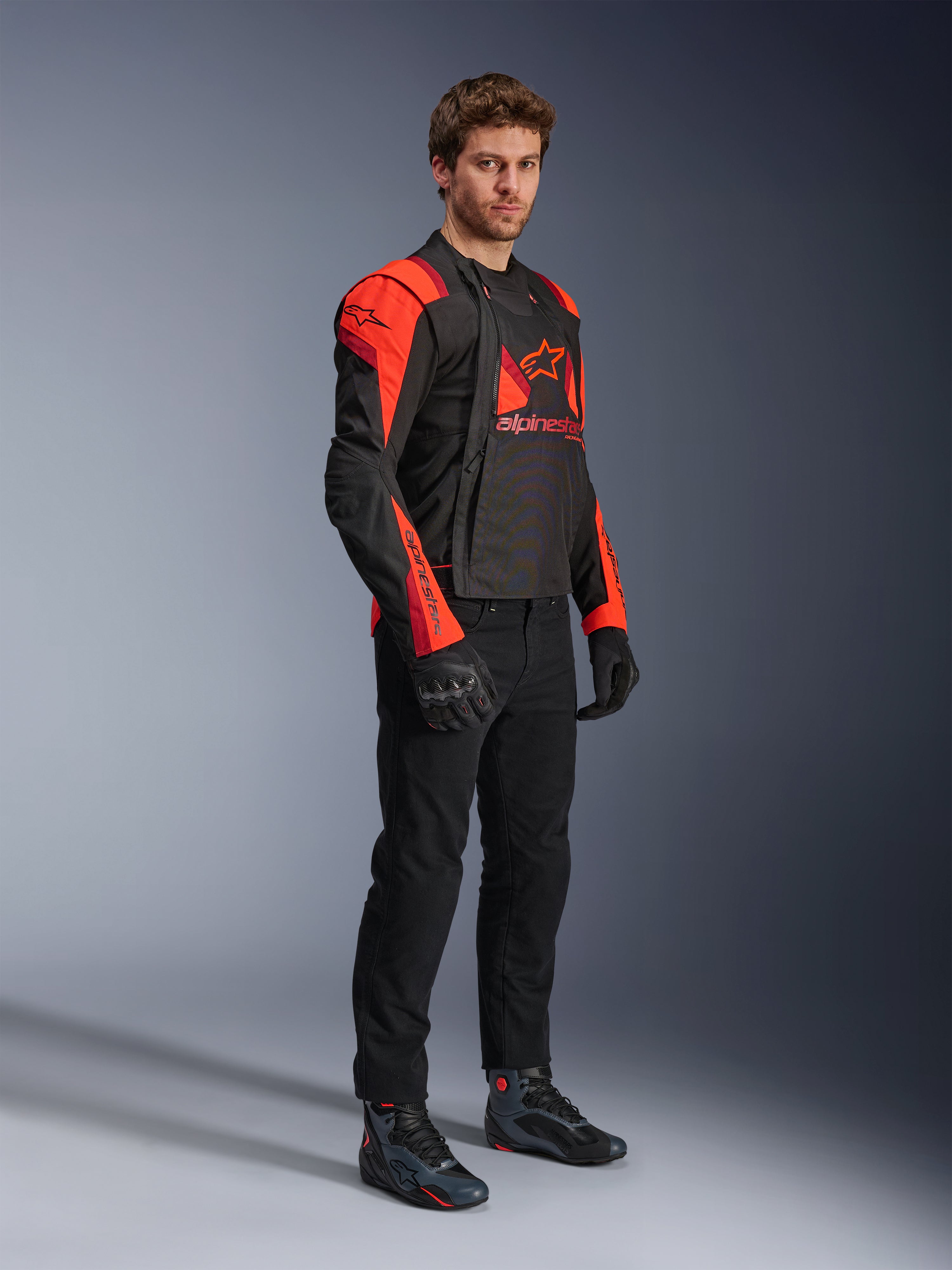 Alpinestars T-Stunt Air Jacket, modulaire motorjas in zwart, Red Fluo en donkerrood, gedragen door een model met geopende voorpanelen die het mesh-interieur onthullen, gecombineerd met een zwarte motorbroek, handschoenen en schoenen.