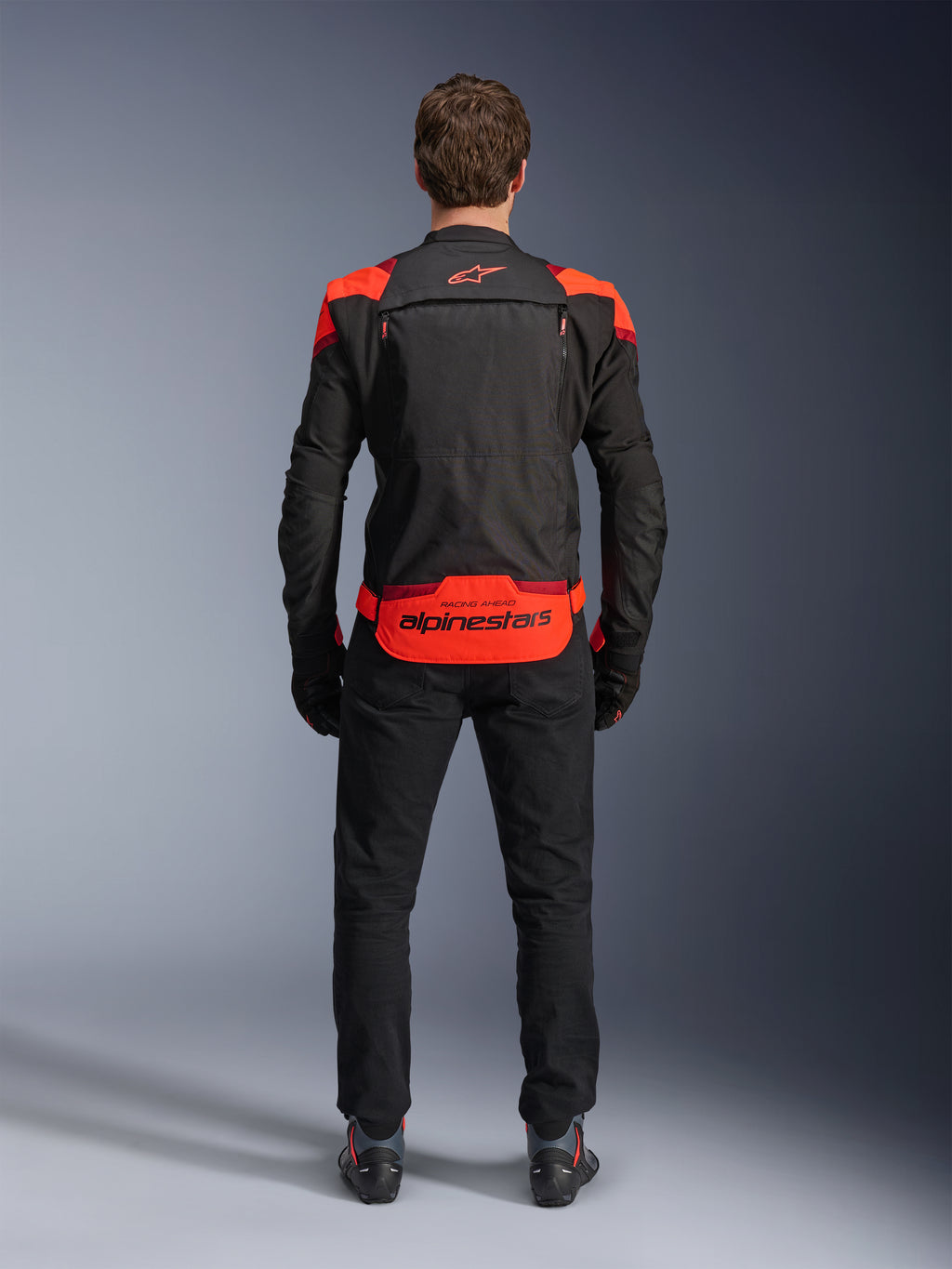 Alpinestars T-Stunt Air Jas, zwart met Red Fluo en Dark Red accenten, achteraanzicht op model, gecombineerd met zwarte motorbroek en laarzen, opvallende Alpinestars-branding op het onderste rugpaneel, modulair ontwerp met mesh-constructie voor ventilatie