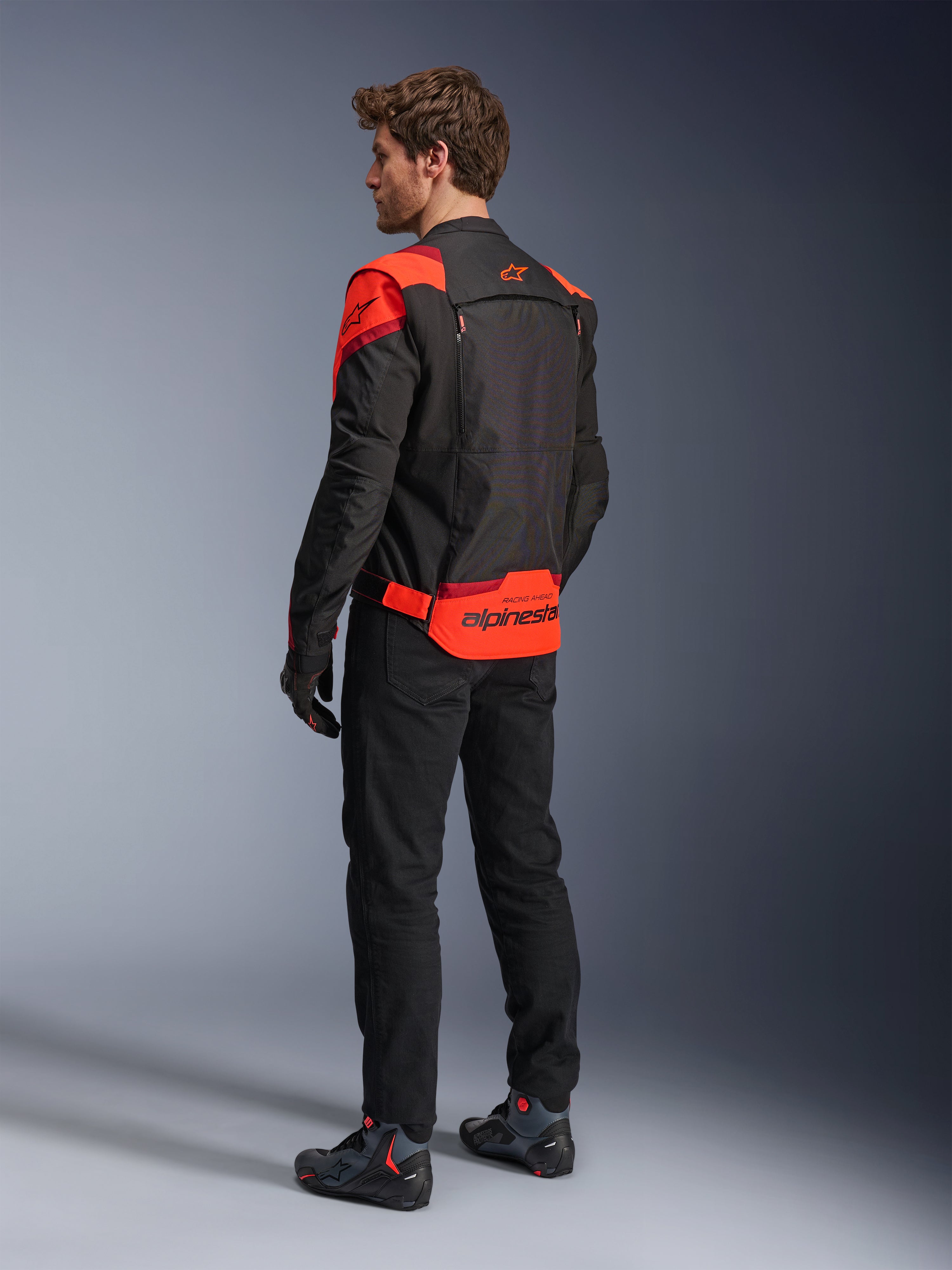 Alpinestars T-Stunt Air Jacket van achteren gezien, zwart, fluorood en donkerrood, textiel motorjas met mesh-panelen en sportieve snit, met opvallende Alpinestars-branding op de onderrug, man draagt de jas met een zwarte broek en motorlaarzen