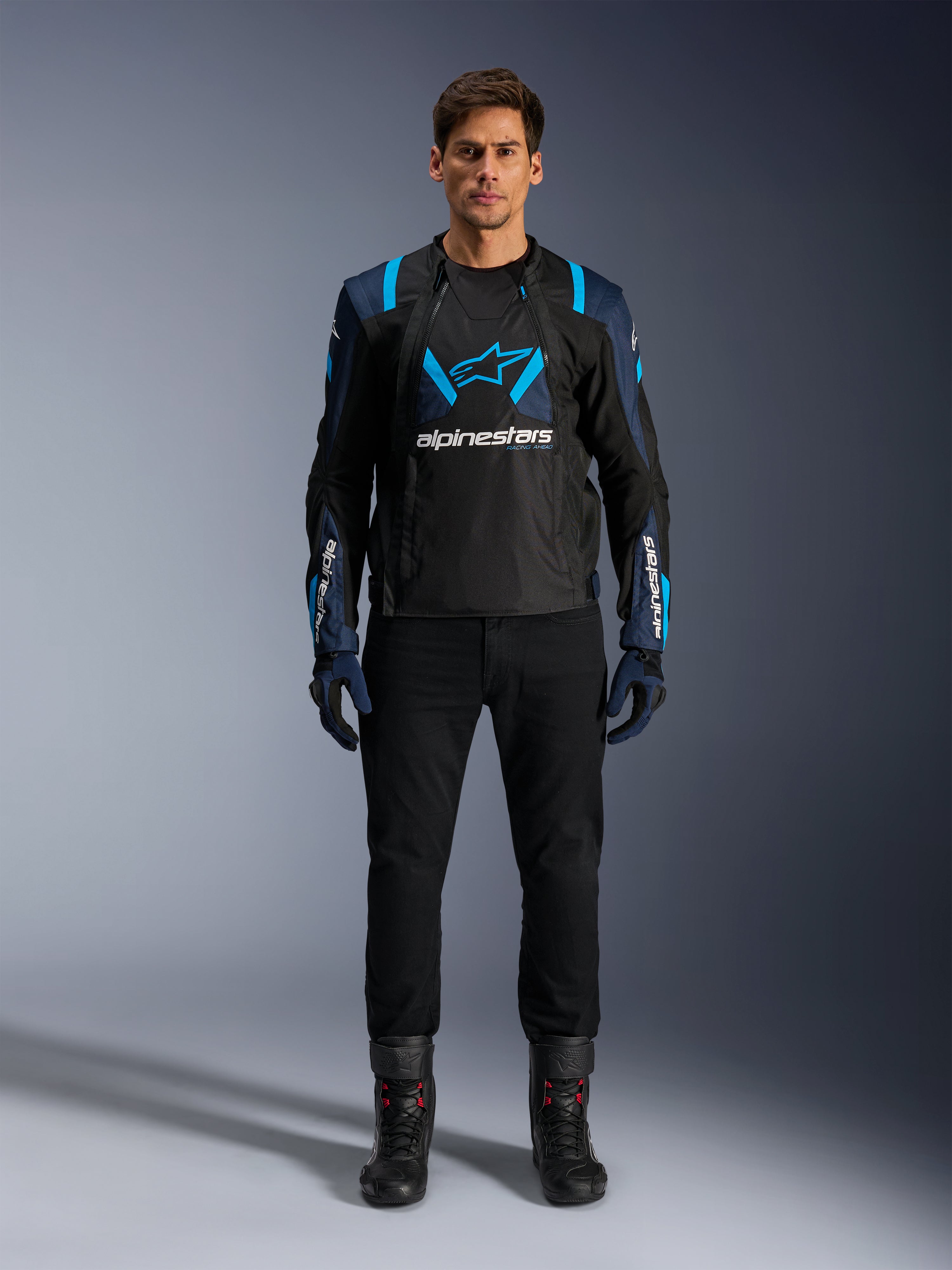 Alpinestars T-Stunt Air jas, motorjas, zwart met donkerblauwe en felblauwe accenten, groot wit logo op de borst, sportief gesneden mesh-constructie voor luchtstroom, gedragen met zwarte motorbroek en laarzen voor een strakke look op de weg