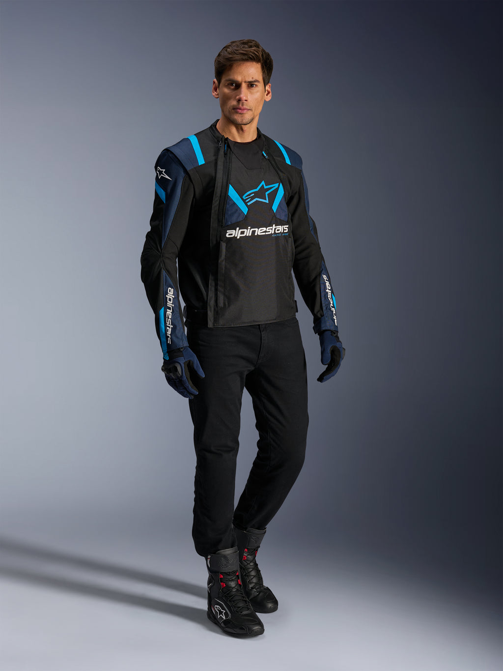 Mannelijk model in Alpinestars T-Stunt Air Jacket, Black Dark Blue en Blithe Blue, met opvallende mesh-panelen voor ventilatie, gecombineerd met een zwarte broek en motorhandschoenen, sportief gesneden motorjas voor zomerse ritten op de weg.