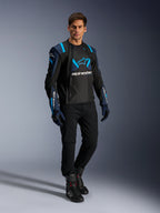 Mannelijk model in Alpinestars T-Stunt Air Jacket, Black Dark Blue en Blithe Blue, met opvallende mesh-panelen voor ventilatie, gecombineerd met een zwarte broek en motorhandschoenen, sportief gesneden motorjas voor zomerse ritten op de weg.
