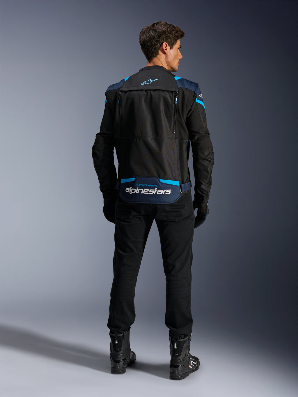 Alpinestars T-Stunt Air Jacket, modulaire textiele motorjas, zwart, donkerblauw en felblauw, achteraanzicht met het opbergvak voor mouwen op de onderrug, witte Alpinestars-branding, gedragen met zwarte broek en laarzen.