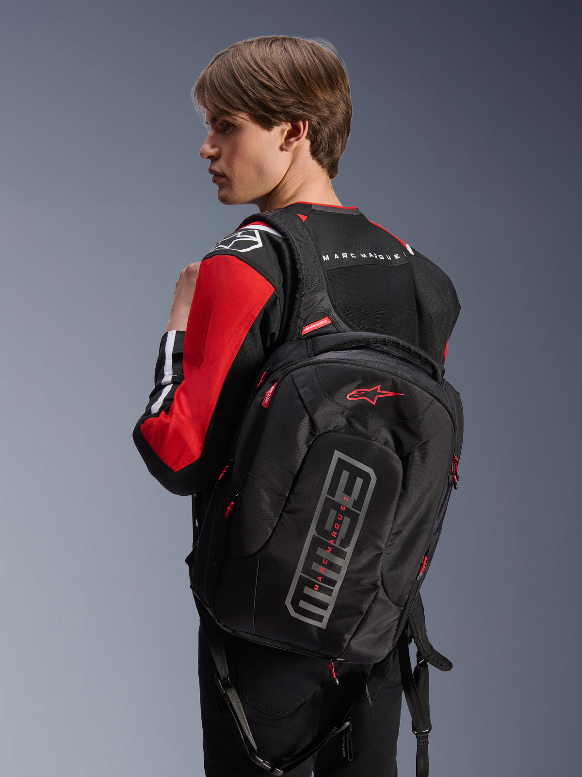 Alpinestars MM93 City Hunter V2 rugzak, zwart, voorzien van rood Astars-logo en grijze MM93-branding, gedragen door een motorrijder in een rood-zwarte motorjas, stedelijke motorkleding uit de Marc Márquez-collectie