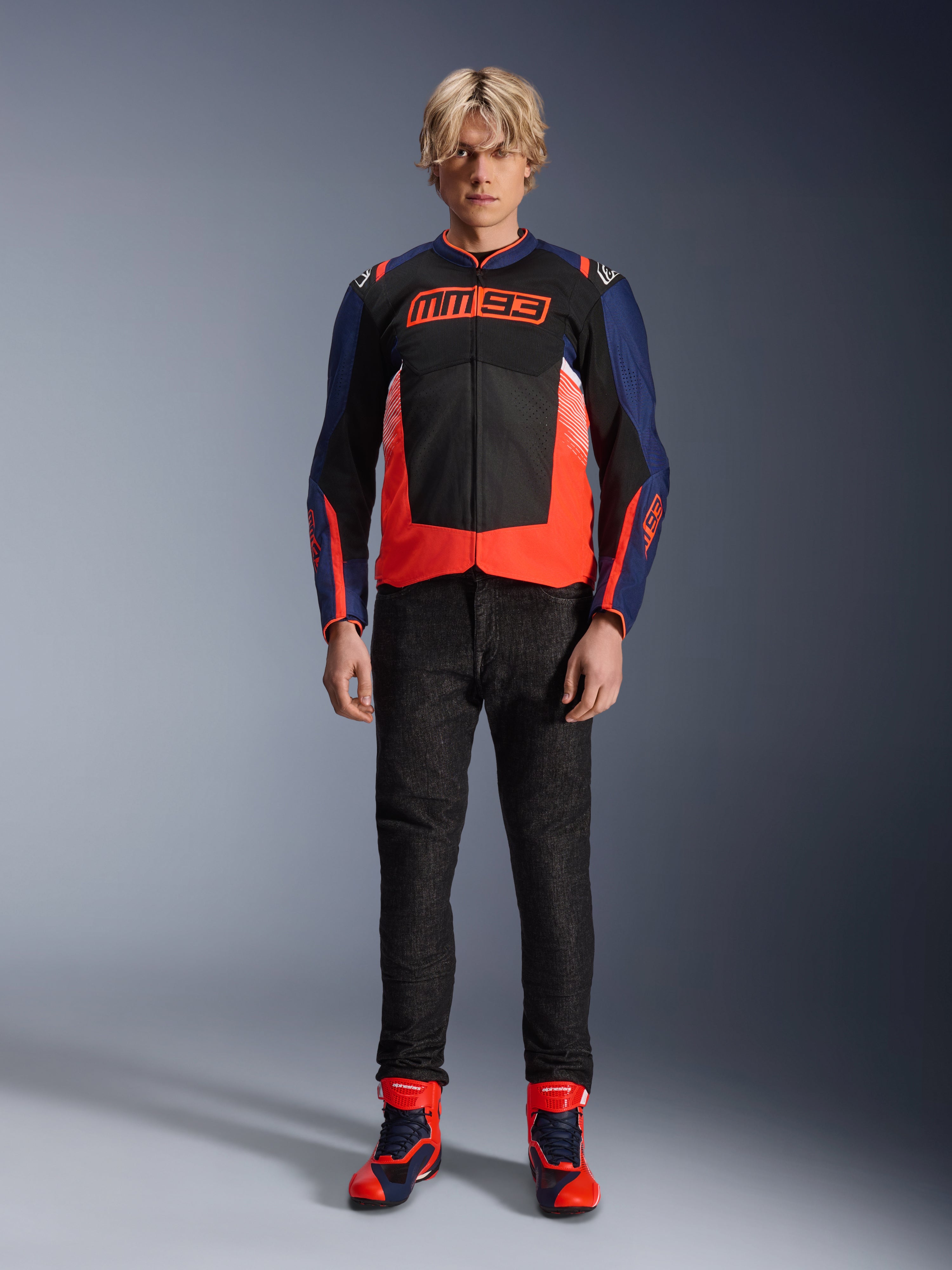 Alpinestars MM93 T-SPS Air V2 Jas, Blue Navy en zwart met Red Fluo accenten, voorzien van het kenmerkende MM93-logo op de borst, gedragen met een zwarte motorbroek en bijpassende blauwe en rode laarzen, stedelijke motoroutfit