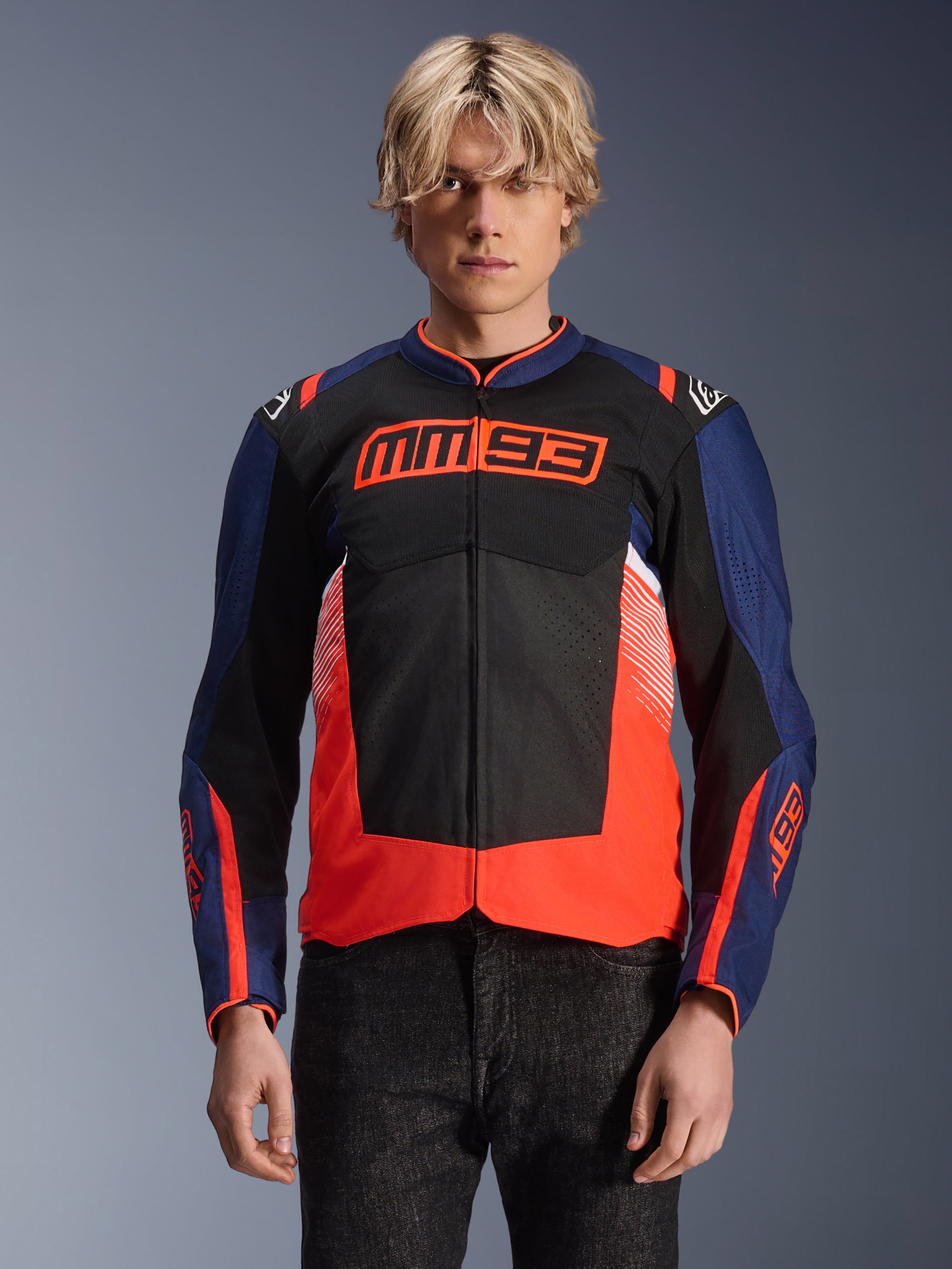Alpinestars MM93 T-SPS Air V2 Jas, textielen motorjas, marineblauw, zwart en fluorescerend rood, gedragen door een persoon, met opvallende MM93-branding op de borst, mesh-panelen voor ventilatie en een modern design met kleurvlakken