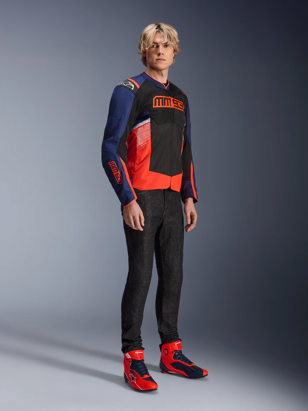 Alpinestars MM93 T-SPS Air v2 Jas in Blue Navy, Black en Red Fluo, met opvallende MM93-branding op de borst en mouwen, gedragen door een mannelijk model met een donkere jeans en bijpassende rood-blauwe motorlaarzen, sportieve urban motoruitrusting