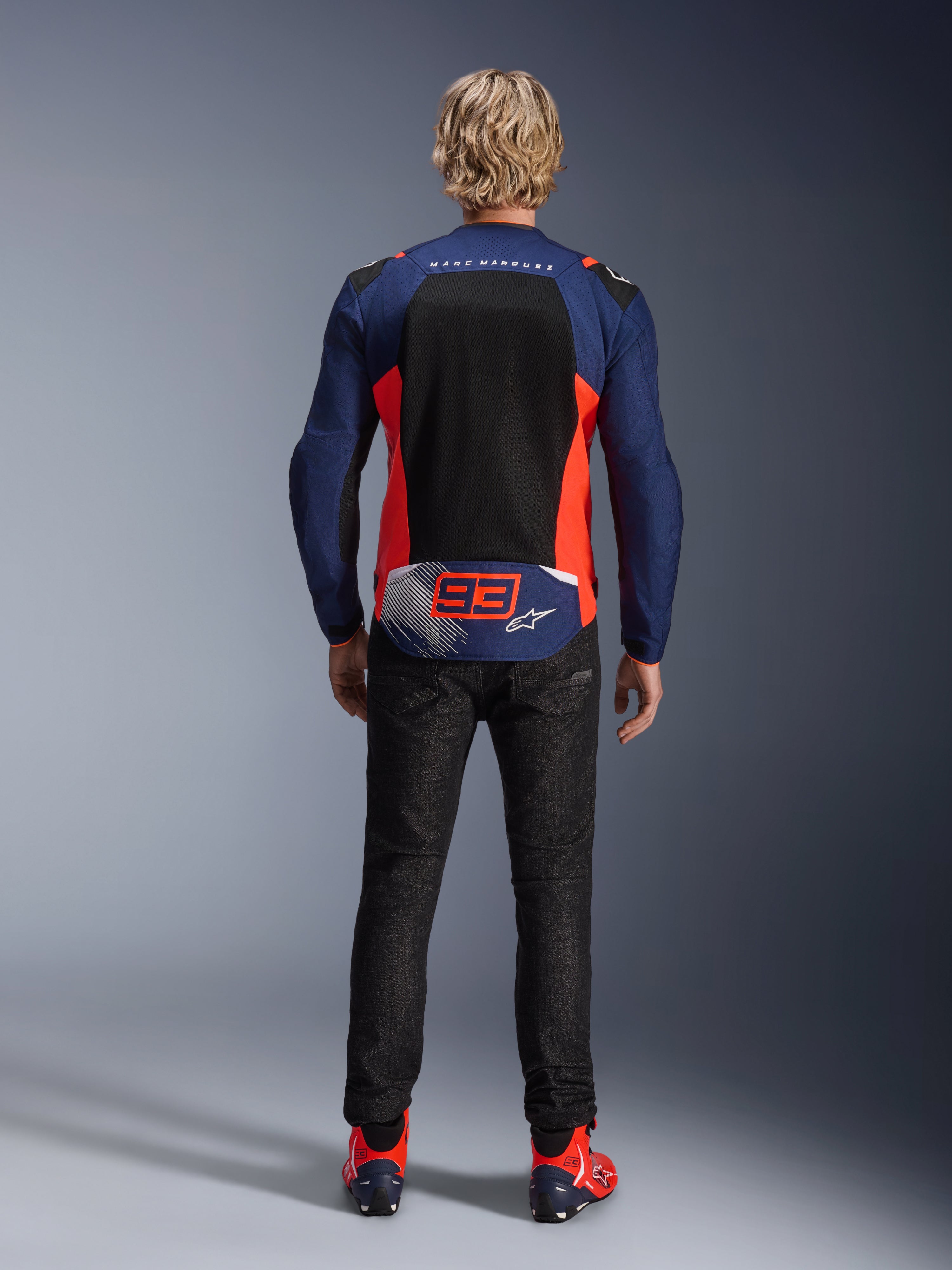 Alpinestars MM93 T-SPS Air V2 Jas, achteraanzicht van een model, Blue Navy met Red Fluo en zwarte accenten, voorzien van een groot mesh rugpaneel en '93' Marc Marquez-branding, gecombineerd met een donkere denim broek en bijpassende motorlaarzen