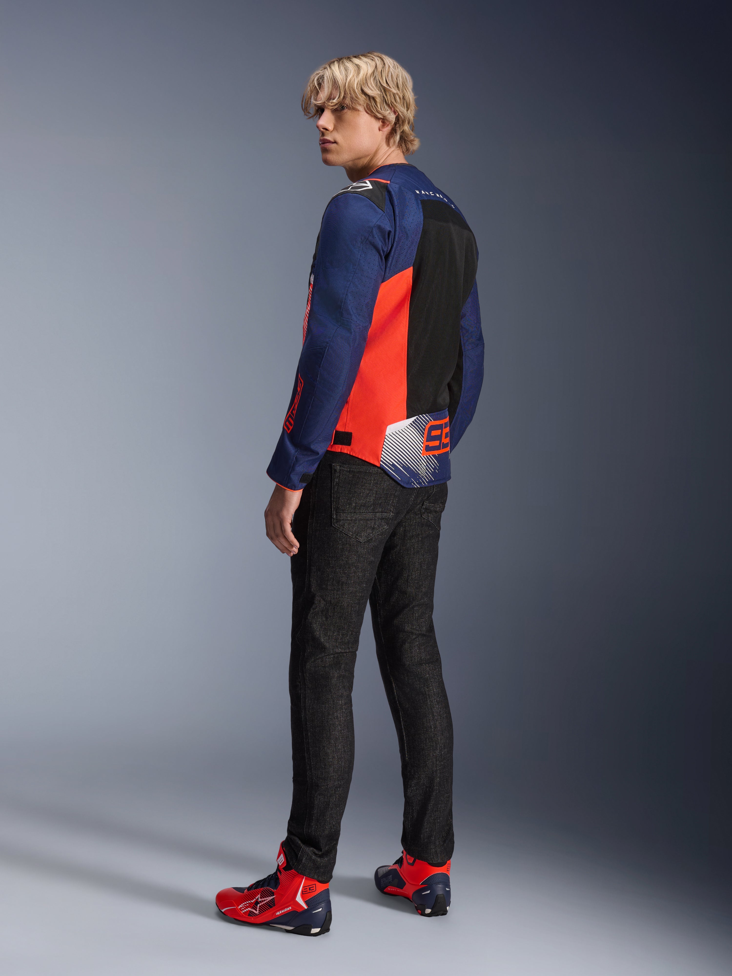 Alpinestars MM93 T-SPS Air V2 Jas, Blue Navy en Red Fluo, gedragen door een model vanaf de achterkant met zwarte motorjeans en bijpassende rood-blauwe schoenen, voorzien van mesh-panelen en Marc Márquez 93 branding.