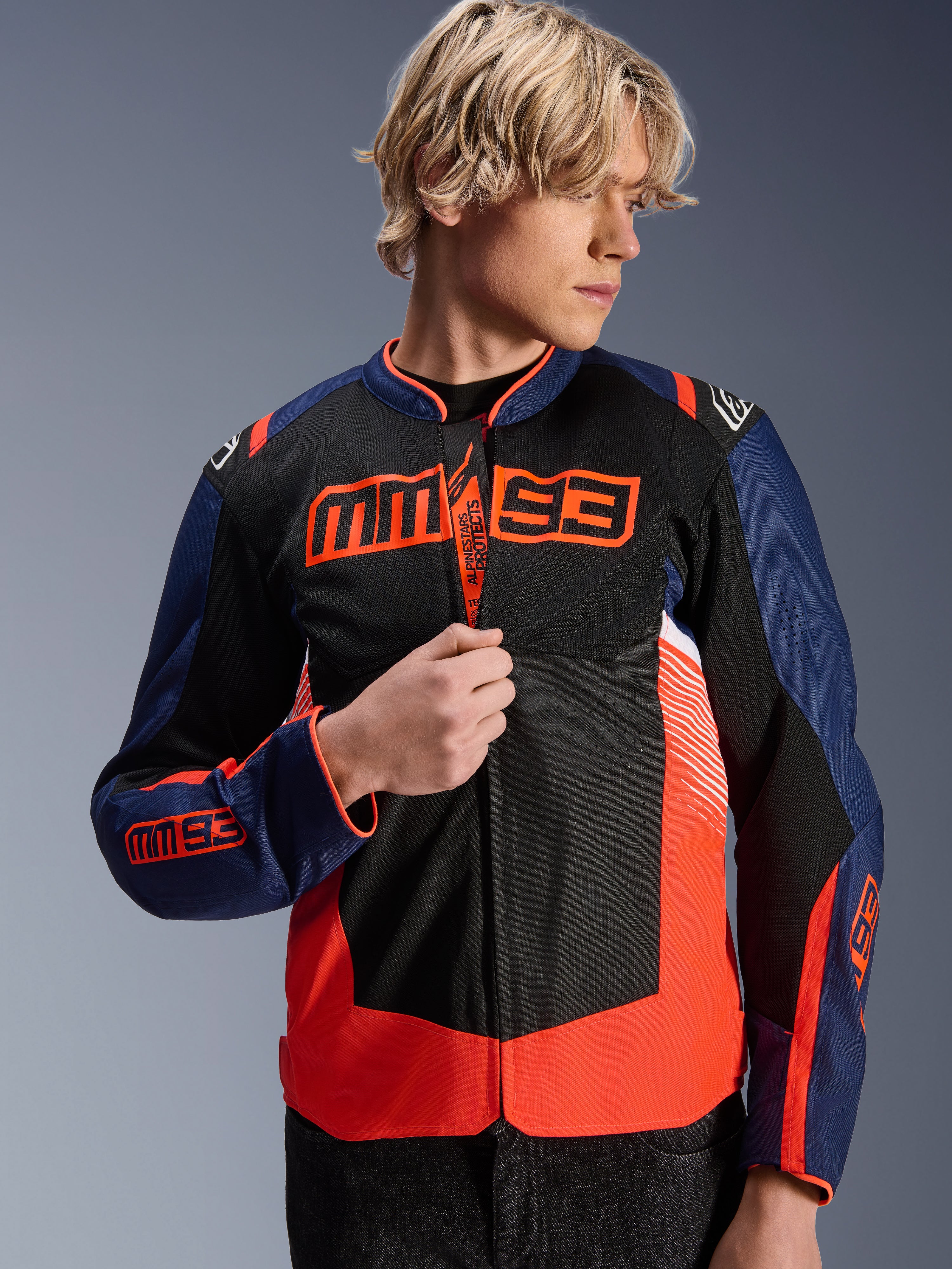 Alpinestars MM93 T-SPS Air V2 Jas, donkerblauwe, zwarte en fluorescerend rode textiele motorjas, gedragen door een mannelijk model dat de rits verstelt, met opvallende Marc Márquez MM93-branding op de borst en mouwen, met geventileerde mesh-panelen voor motorrijden op de weg in de zomer.