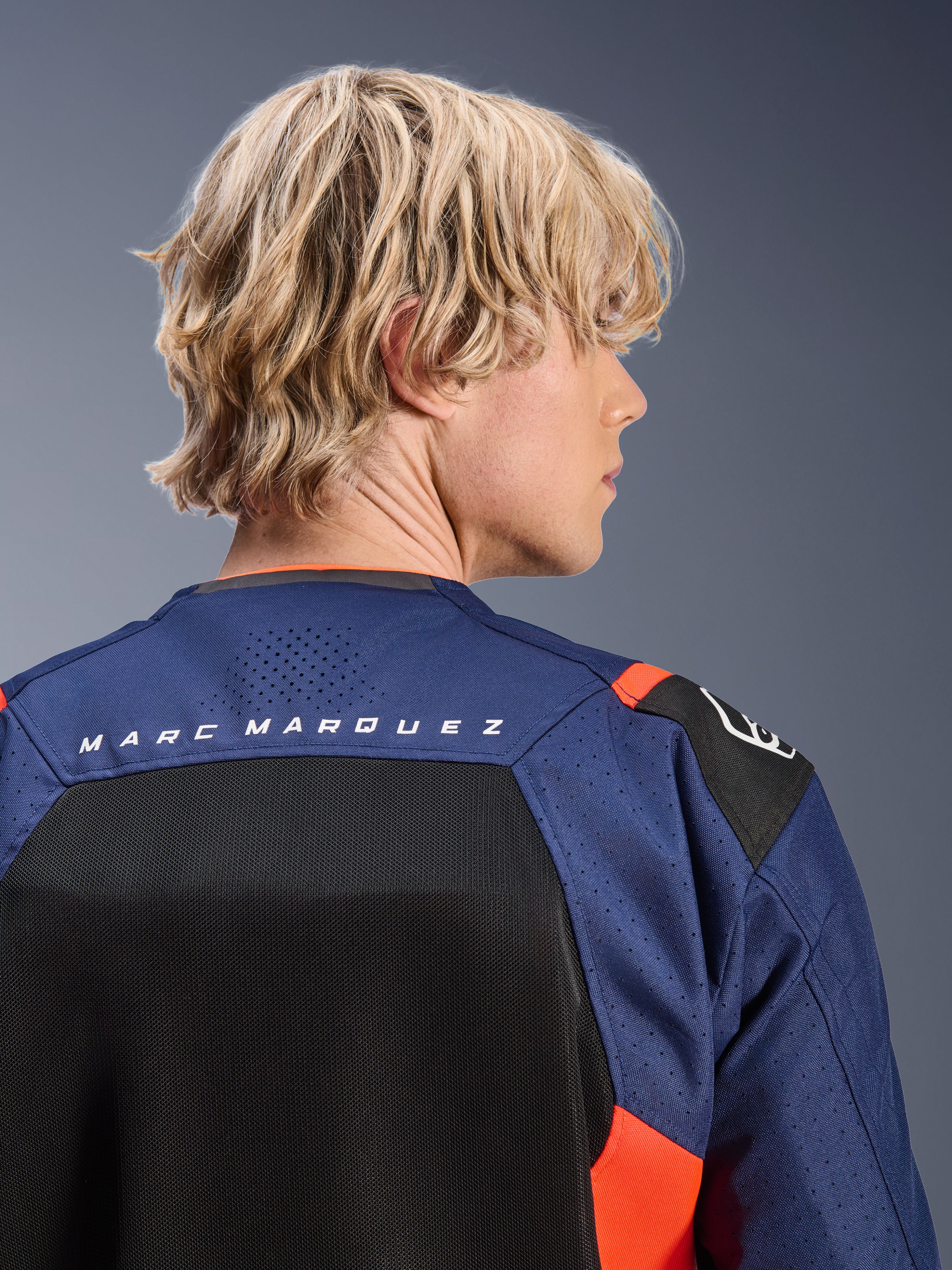 Alpinestars MM93 T-SPS Air V2 Jas, Blue Navy en zwart met Red Fluo accenten, close-up van de bovenrug met 'MARC MARQUEZ' branding, ademende mesh-panelen en geperforeerde textielconstructie, gedragen door een blond model