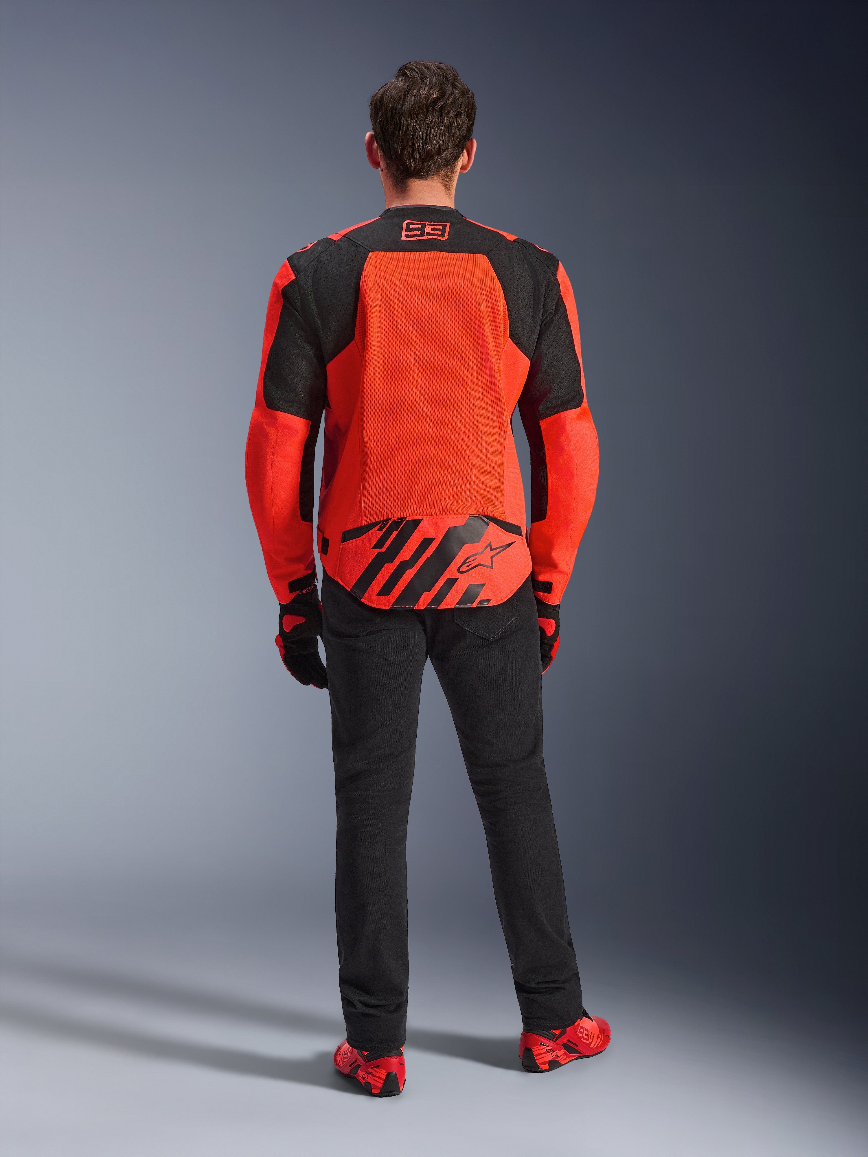 Achteraanzicht van de MM93 T-SPS Air V2 Jas, Red Fluo en Black, textielen motorjas met grote mesh-panelen op de rug voor ventilatie, gedragen met zwarte broek en rood schoeisel, voorzien van Marc Marquez 93-branding