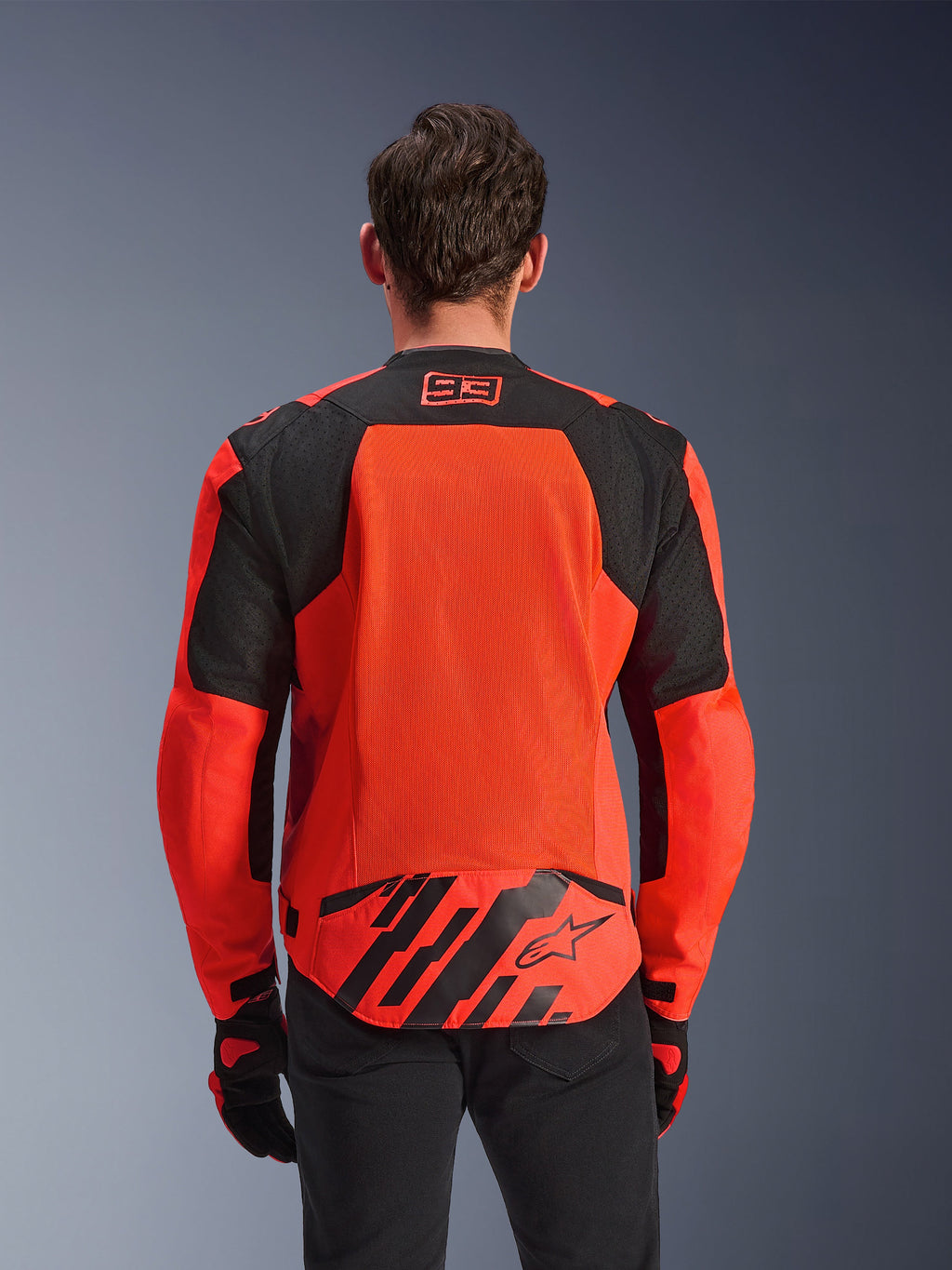 Alpinestars MM93 T-SPS Air V2 Jas, achteraanzicht, fluo rood en zwart, met 93-racelogo op de nek, groot mesh-paneel op de rug voor luchtstroom, achterflap onderaan met Alpinestars-sterlogo en geometrische strepen, gedragen met een zwarte broek en motorhandschoenen