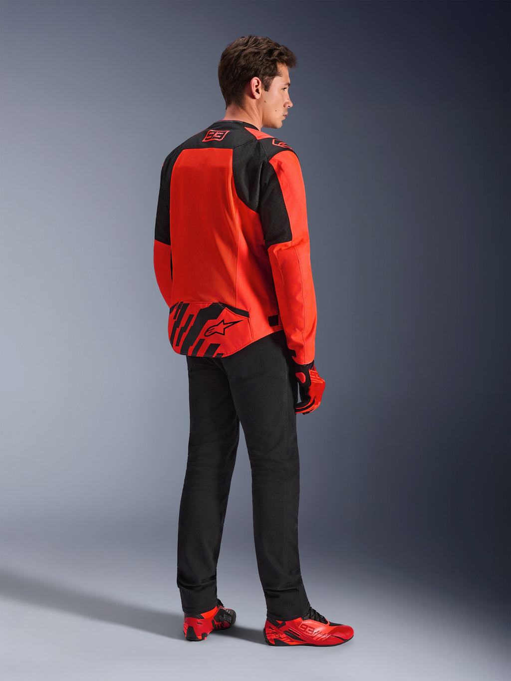 Alpinestars MM93 T-SPS Air V2 Jas in Red Fluo en Black, driekwart achteraanzicht op een model, met Marc Marquez 93-branding op de kraag en onderrug, gecombineerd met een zwarte broek en rode motorschoenen.