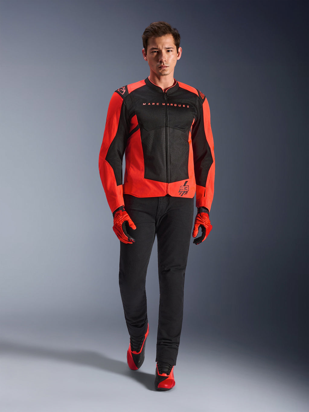 Alpinestars MM93 T-SPS Air V2 Jas, fluo rood en zwart, gedragen door een model met bijpassende rode handschoenen en zwarte broek, voorzien van Marc Marquez-branding en het 93-logo, geventileerde sportieve motorjas voor stadsritten