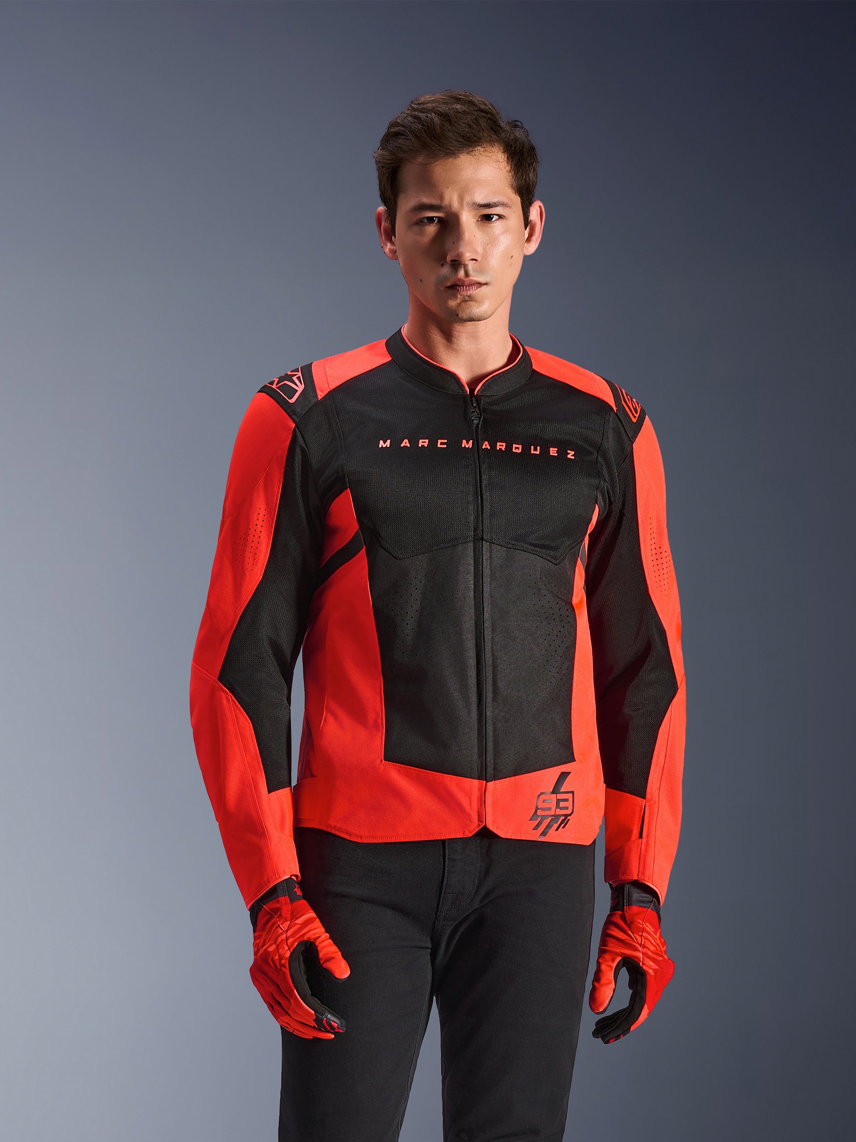 Alpinestars MM93 T-SPS Air V2 Jas, motorjas, fluo rood en zwart, gedragen door een model met bijpassende rode handschoenen en zwarte broek, met 'Marc Marquez' branding op de borst en '93' logo, sportieve mesh-constructie