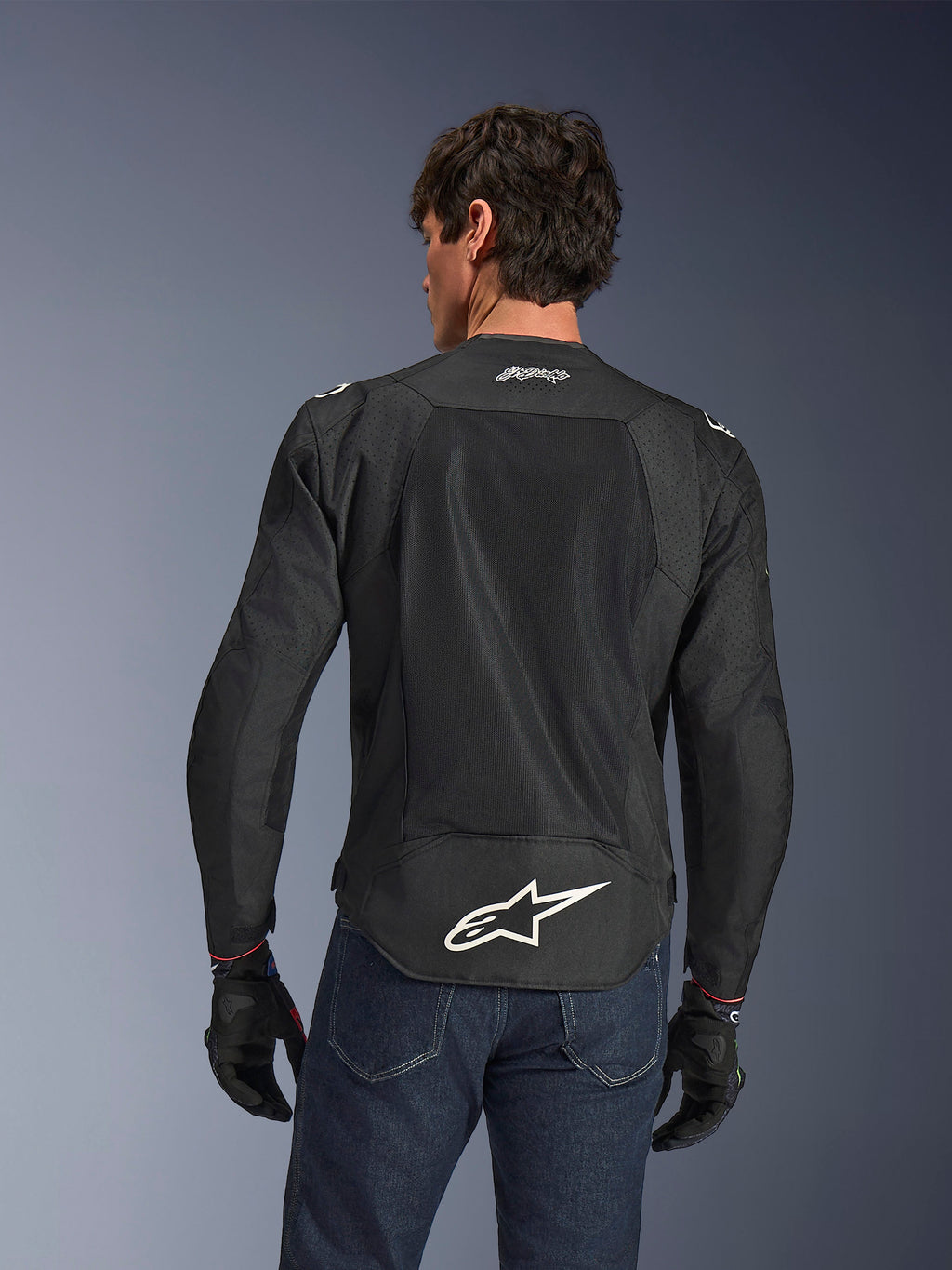 Alpinestars FQ20 T-SPS Air V2 Monster Jacket, Black, Rear view featuring large ventilation mesh panels and white Alpinestars logo on the lower back, worn with blue jeans and black motorcycle gloves, urban sport riding style.