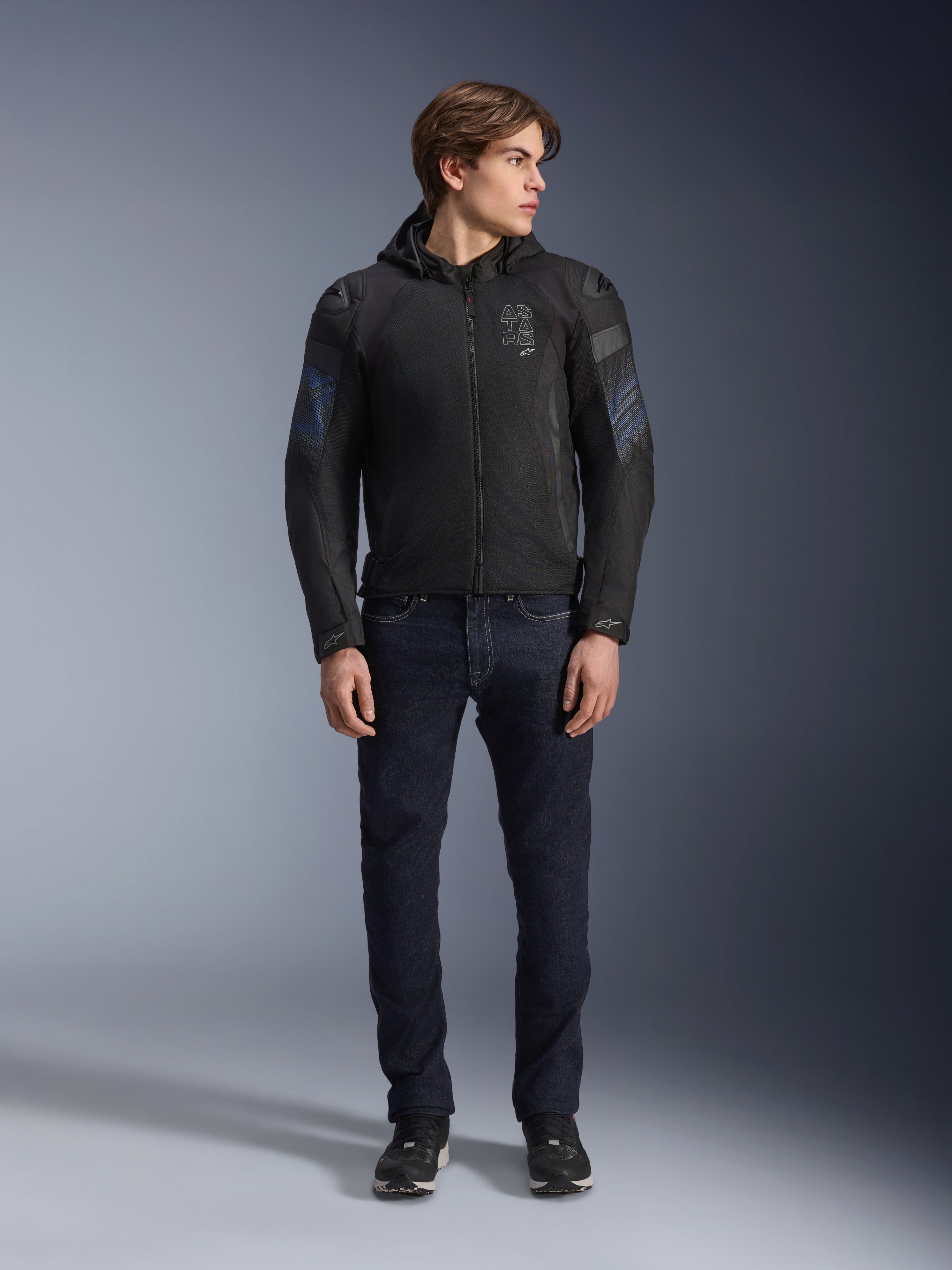 Mannelijk model draagt de Alpinestars Zaca Air Venom WP jas in zwart en elektrisch blauw, textiele motorjas met 3D-mesh panelen en een afneembare capuchon, gecombineerd met een donkere denim jeans, moderne sportgeïnspireerde motoruitrusting