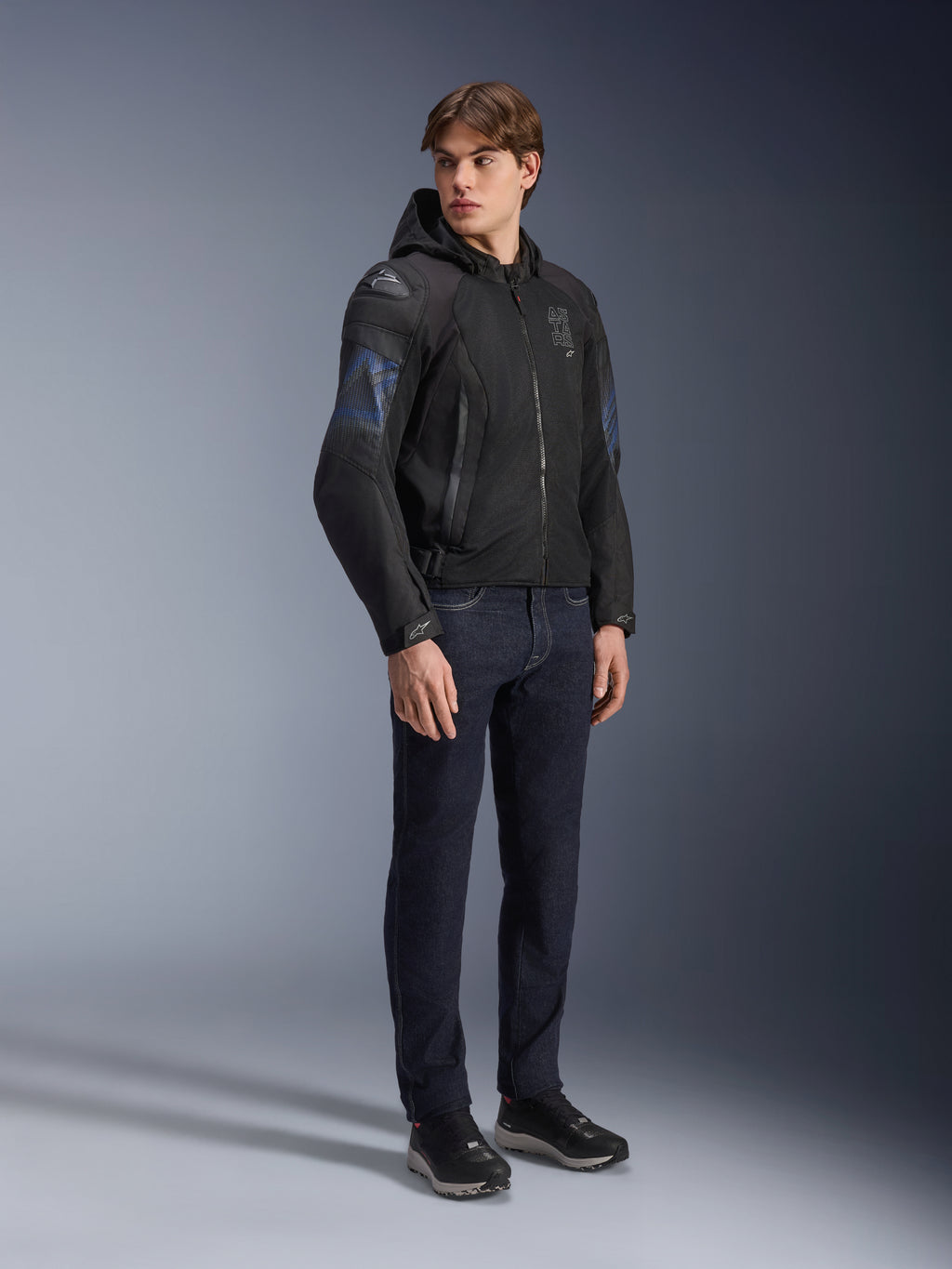 Alpinestars Zaca Air Venom WP Jas, zwarte en elektrisch blauwe textiele motorjas, gedragen door model met donkere jeans en zwarte sneakers, voorzien van een afneembare capuchon, mesh ventilatiepanelen en DFS Lite schouderschuivers, sportieve rijuitrusting voor alle weersomstandigheden.