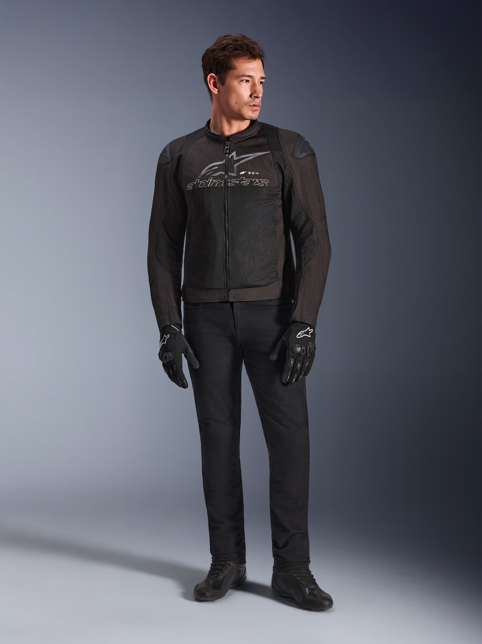 Mannelijk model draagt de Alpinestars SMX Air Jacket, een Black Anthracite motorjas met geventileerde mesh-panelen en een grijs logo op de borst, gecombineerd met een zwarte broek en beschermende handschoenen, studio-opname met een gradiënt achtergrond