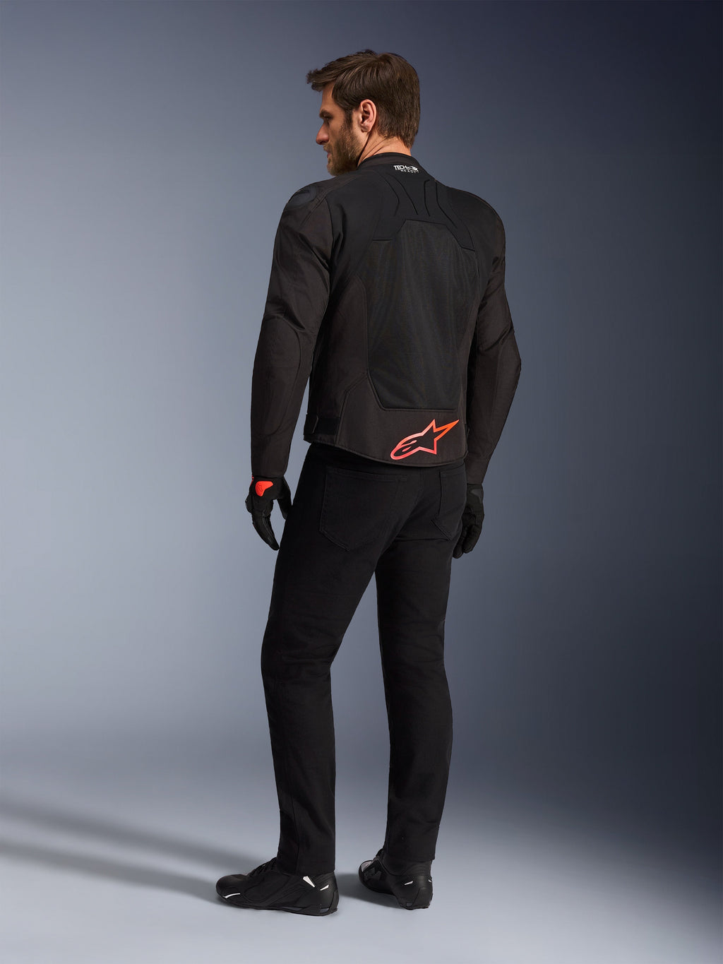 Achteraanzicht van een model die de Alpinestars SMX Air Jacket in het zwart draagt, met een felrood Alpinestars-logo op de onderrug, 'Tech-Air Ready'-branding op de kraag en technische mesh-panelen voor ventilatie, gecombineerd met een zwarte motorbroek en handschoenen.