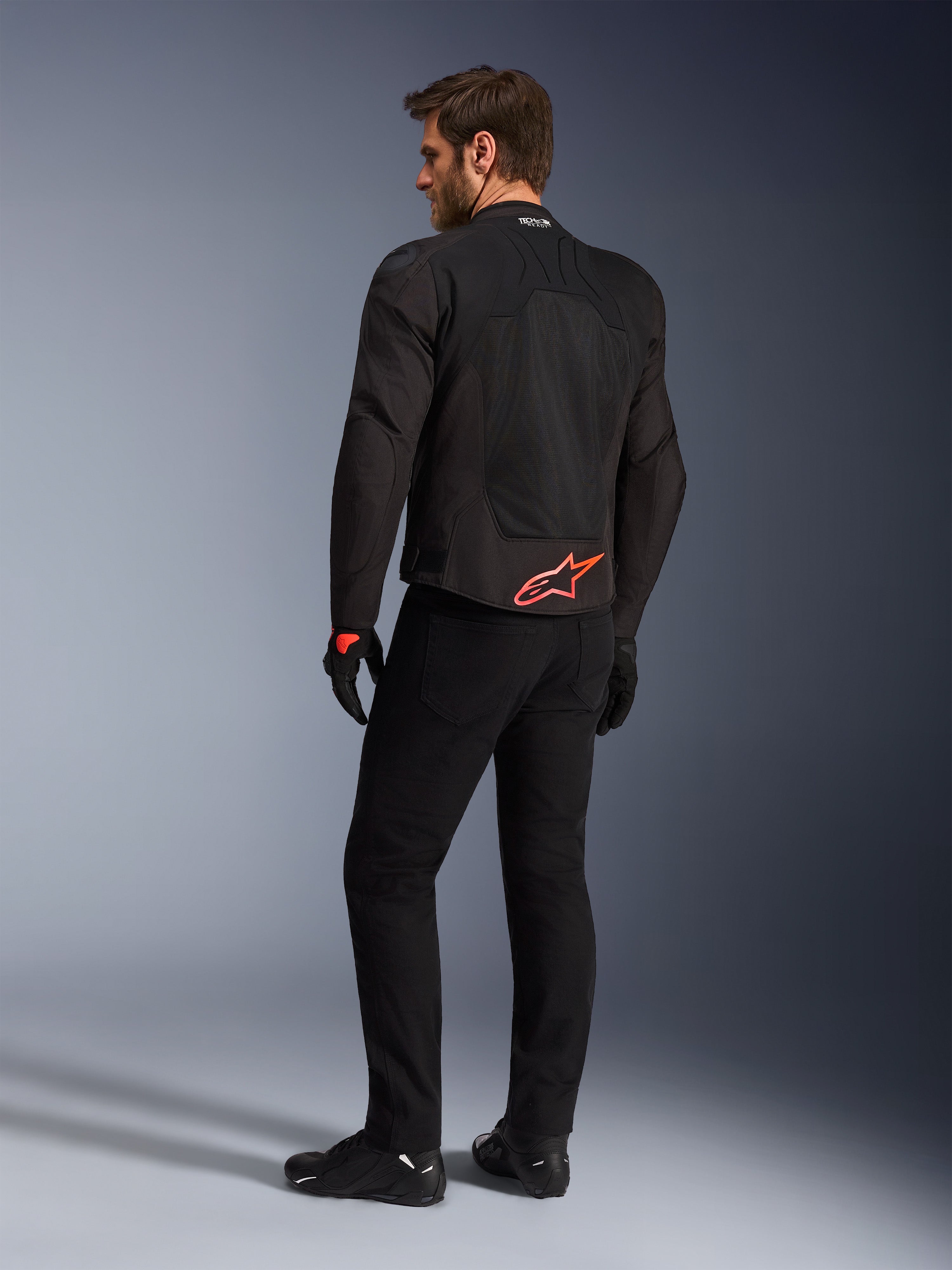 Achteraanzicht van een model die de Alpinestars SMX Air Jacket in het zwart draagt, met een felrood Alpinestars-logo op de onderrug, 'Tech-Air Ready'-branding op de kraag en technische mesh-panelen voor ventilatie, gecombineerd met een zwarte motorbroek en handschoenen.