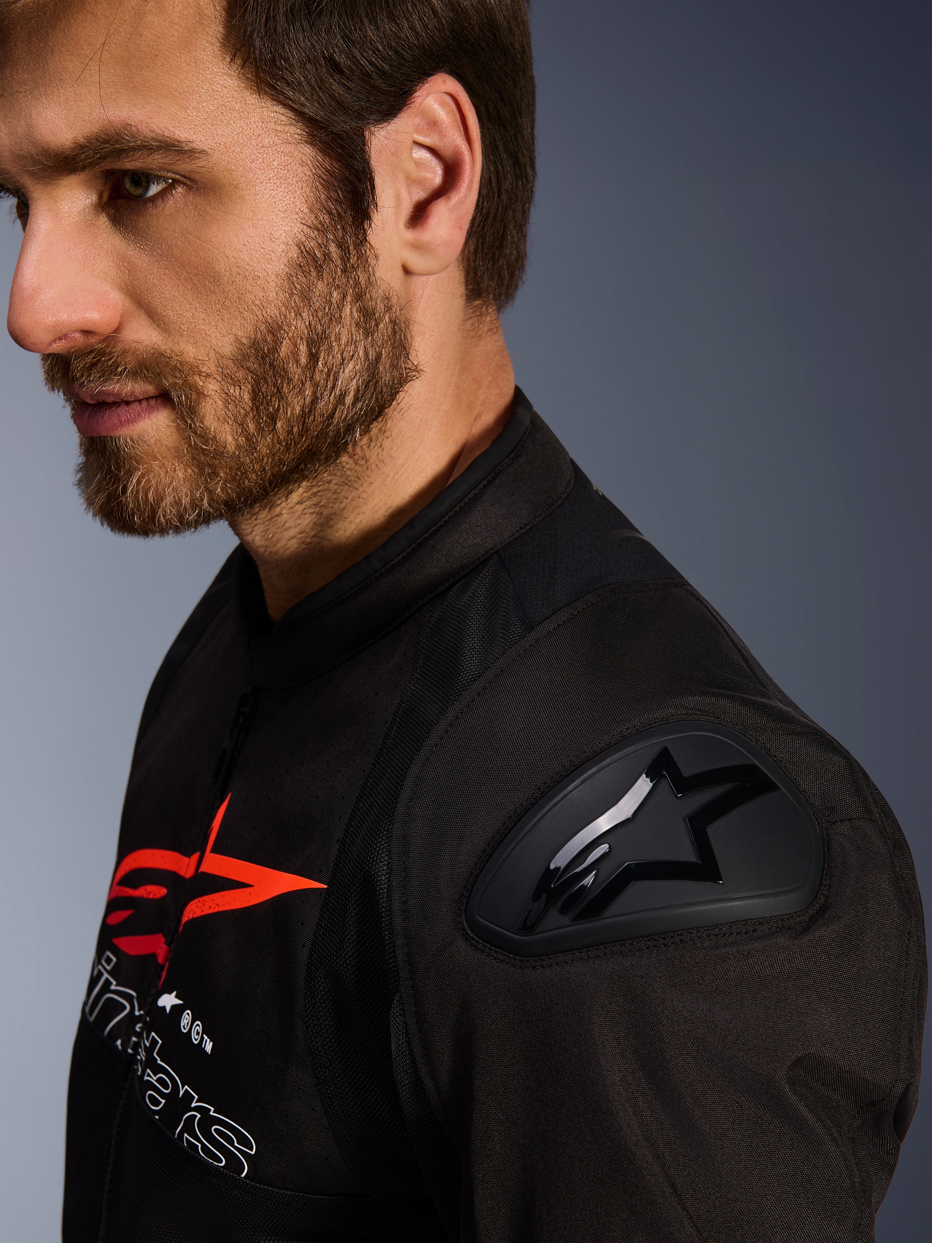 Alpinestars SMX Air Jacket, zwart en rood, close-up detail van de DFS Lite schouderbeschermer met sterlogo, gedragen door een man met een baard, textiele race- en sportmotorjas.