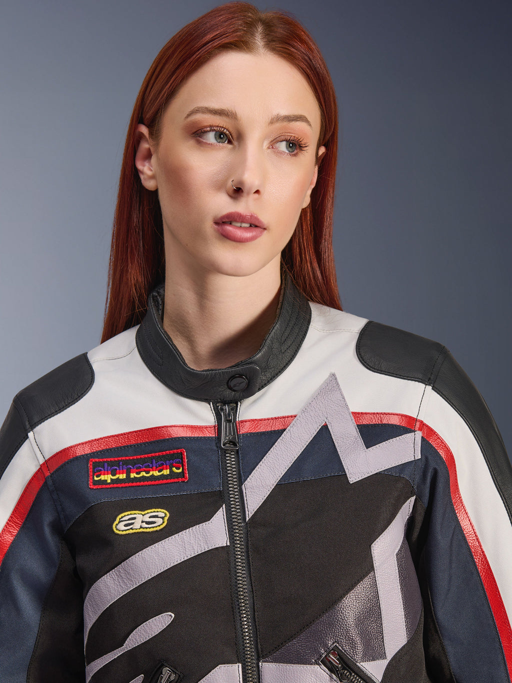 Alpinestars Idles damesjas, close-up van een vrouwelijk model met rood haar, Off White Flame Red en Black kleurstelling, vintage-geïnspireerd racing-ontwerp met logopatches en grijze geometrische ster-graphics, urban motorrijuitrusting.