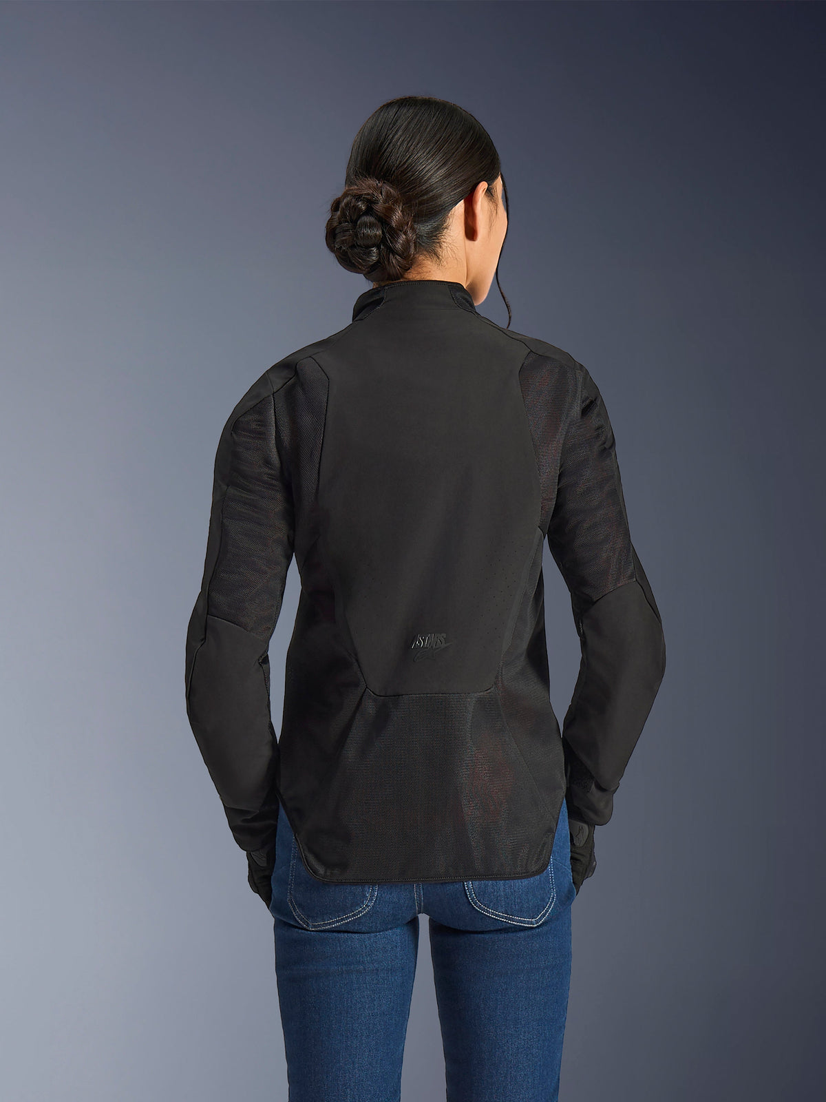 Alpinestars Stella Aeroshell Airflow motorjas voor dames, Black Anthracite, achteraanzicht met geïntegreerde mesh-ventilatiepanelen op de rug en mouwen, lasergeperforeerd softshell-chassis met een verlengd achterprofiel, gedragen met een blauwe denim jeans.