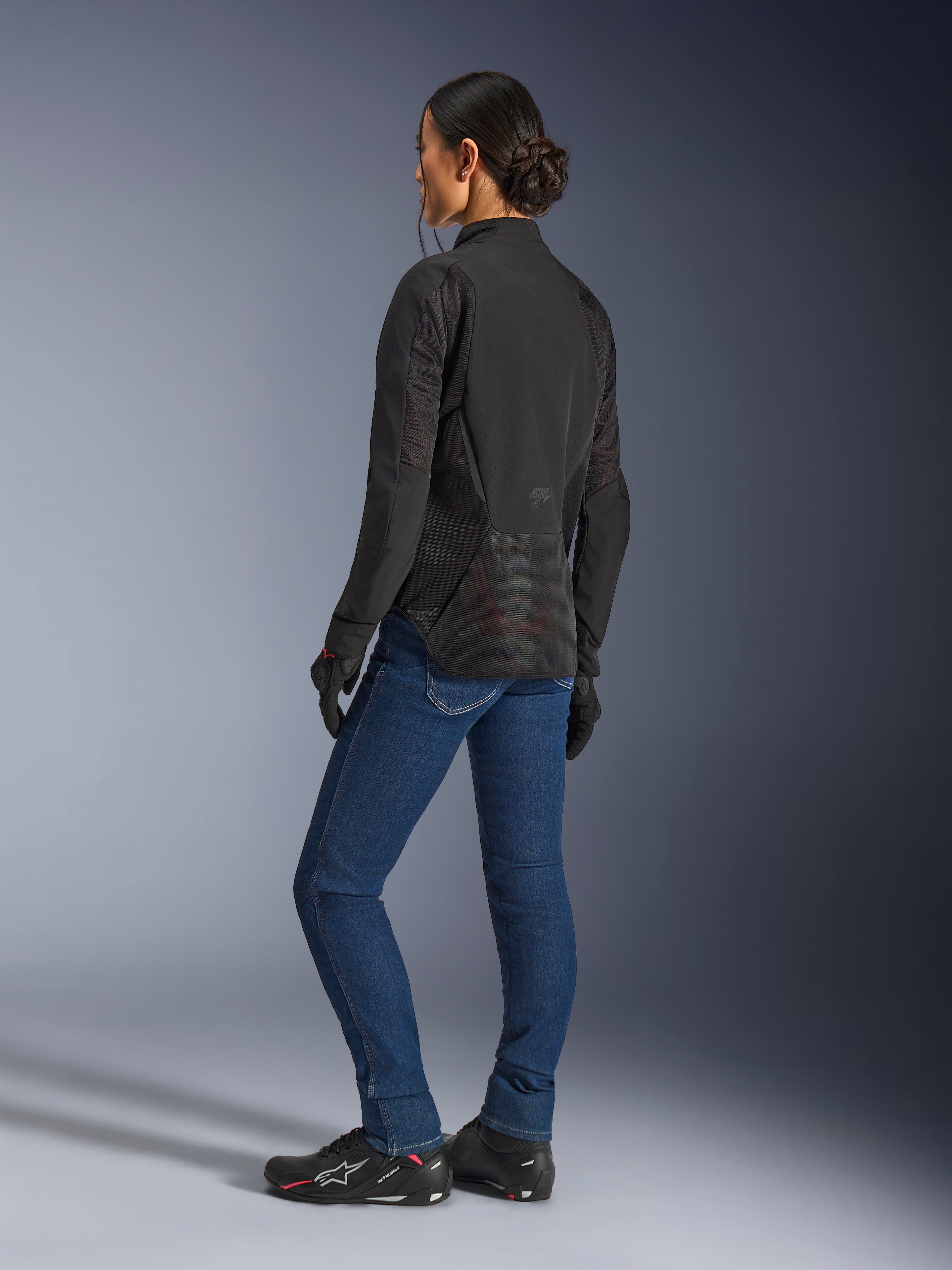 Vrouw draagt de Alpinestars Stella Aeroshell Airflow jas, Black Anthracite, driekwart achteraanzicht met ademende mesh-panelen en een verlengd achterprofiel, gecombineerd met een blauwe denim motorjeans en zwarte Alpinestars-schoenen