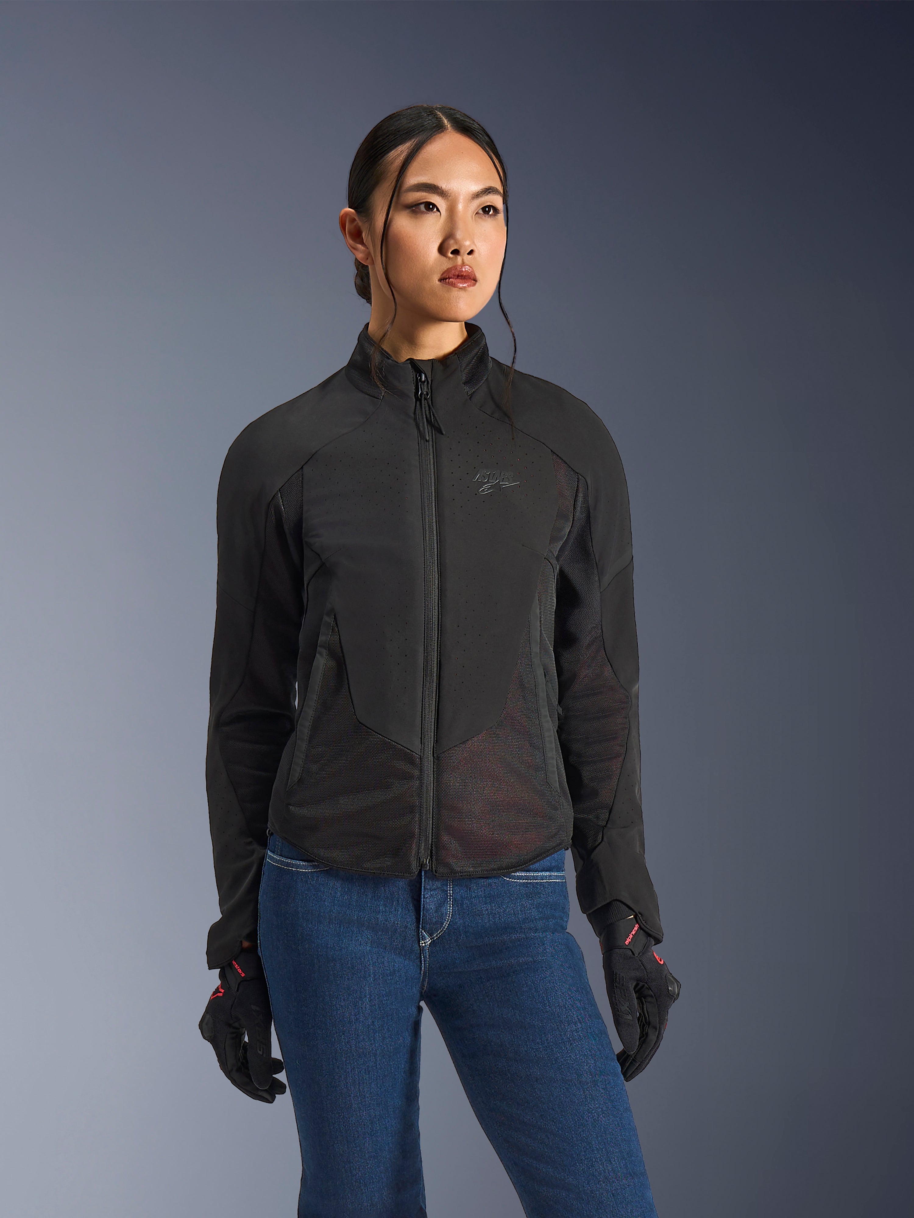 Alpinestars Stella Aeroshell Airflow Jacket, motorjas voor dames, zwart antraciet, gedragen door model met blauwe jeans en zwarte handschoenen, voorzien van laser-geperforeerde softshell en mesh panelen voor optimale ventilatie, ergonomisch sportief ontwerp.