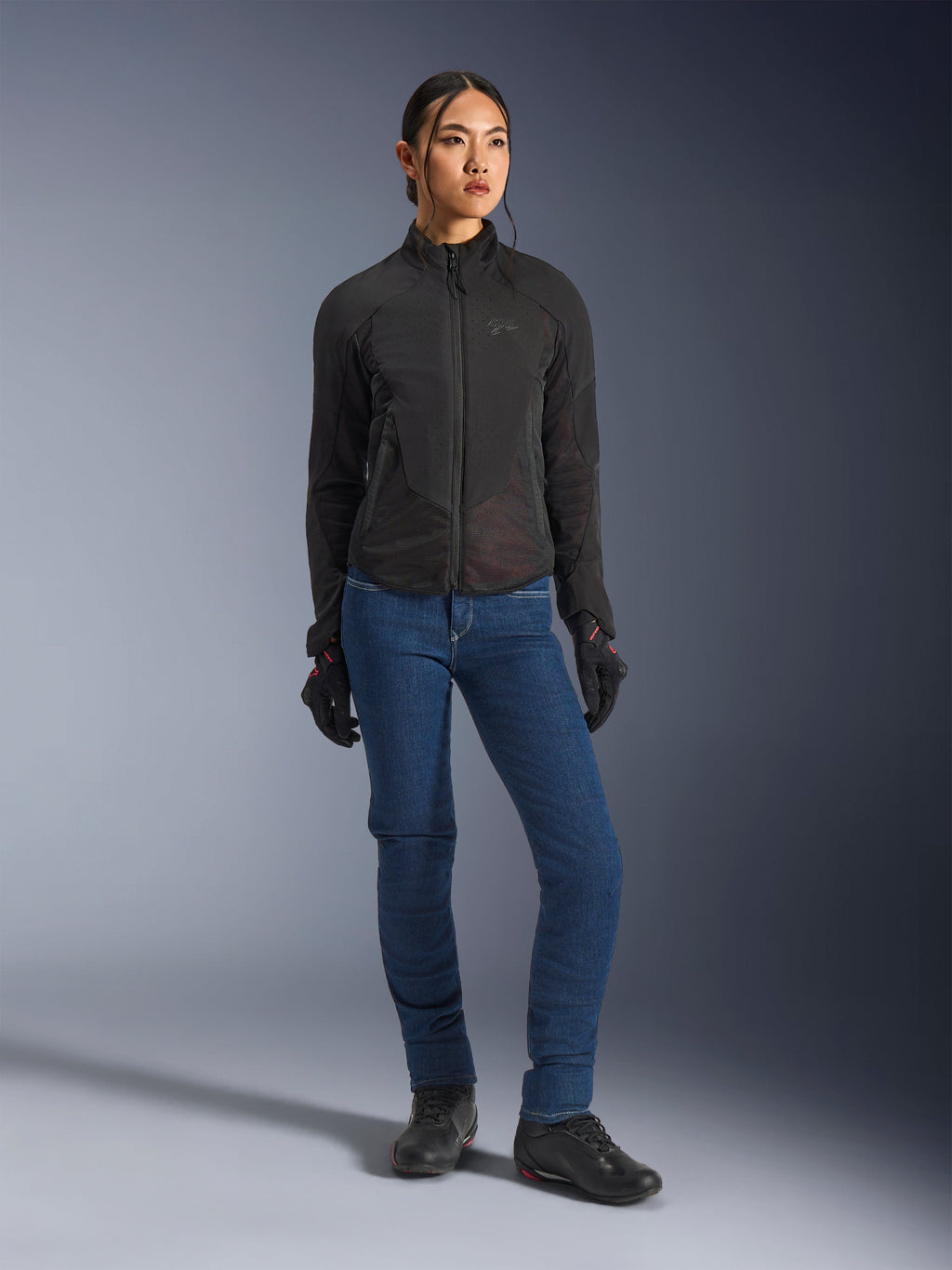 Vrouwelijke motorrijder draagt de Alpinestars Stella Aeroshell Airflow jas in het zwart, voorzien van lasergeperforeerde ventilatie en mesh-panelen, gecombineerd met een blauwe motorjeans en zwarte motoruitrusting, moderne sportieve en urban esthetiek