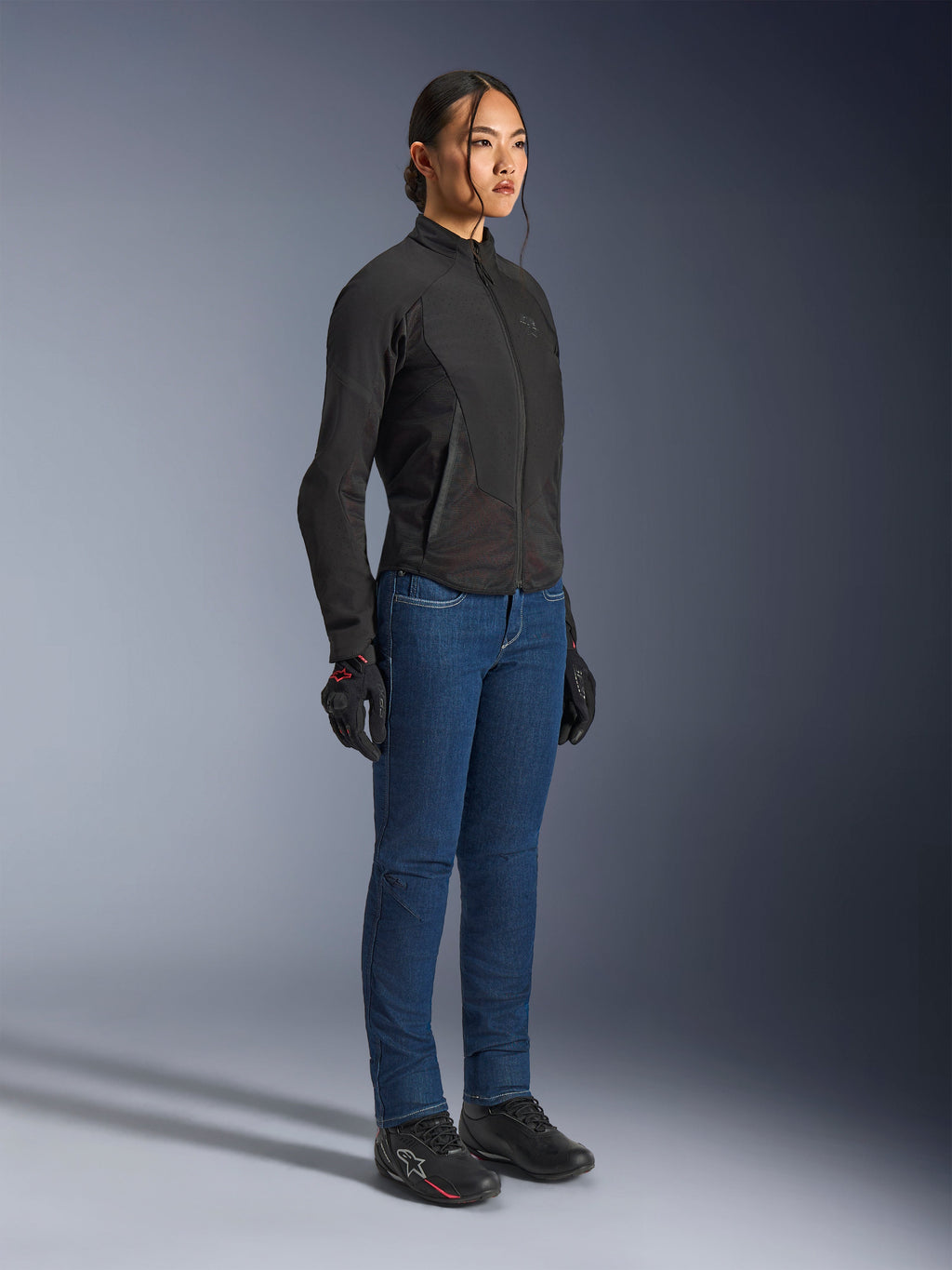 Alpinestars Stella Aeroshell Airflow Jas in Black Anthracite, slim-fit softshell motorjas voor dames, gedragen door een vrouwelijke rijder met blauwe motorjeans en zwarte handschoenen, volledige studio-opname