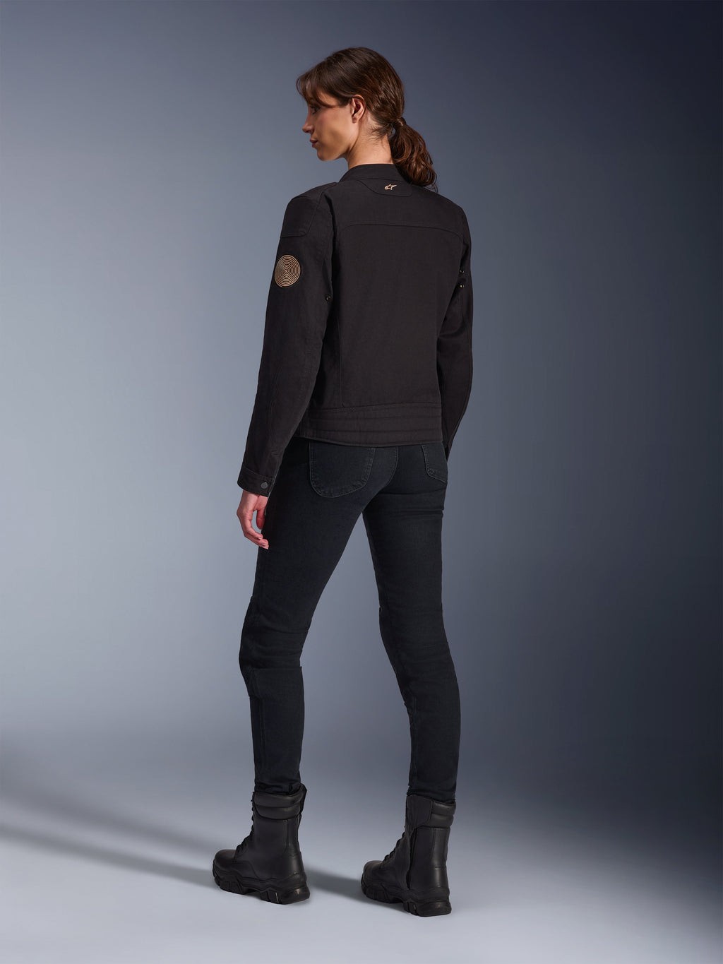 Model draagt de Alpinestars Stella Grace Jas in het zwart, achteraanzicht met de vintage Oscar spiraalpatch op de mouw en een klein sterlogo op de kraag, gecombineerd met een zwarte motorbroek en zwarte laarzen voor stedelijk rijden