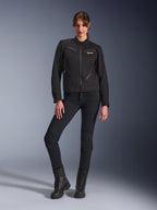 Vrouw in de zwarte Alpinestars Stella Grace Jacket, urban motorjas met werkkleding-esthetiek, voorzien van ritszakken aan de voorzijde en logo op de borst, gecombineerd met een zwarte slim-fit broek en zwarte laarzen, moderne motorkleding voor dames