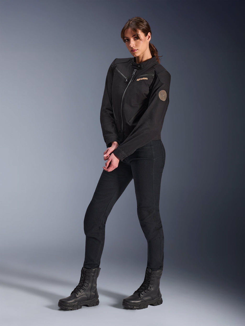 Vrouwelijk model draagt de Alpinestars Stella Grace damesjas, zwarte urban motorjas, gestyled met een zwarte motorbroek en laarzen, op werkkleding geïnspireerd ontwerp met een brons geborduurd logo op de borst en een ronde spiraalvormige patch op de mouw