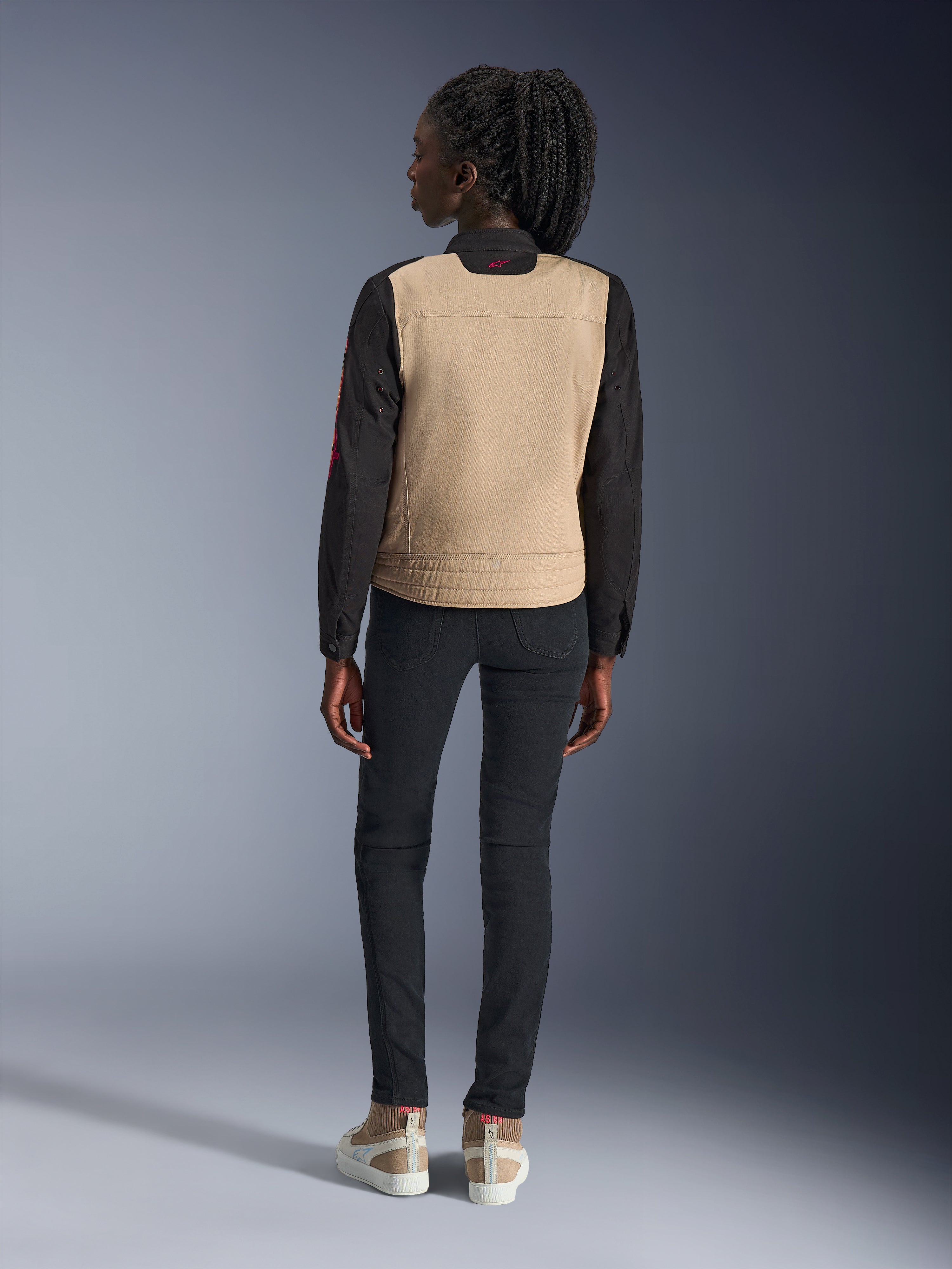 Vrouw draagt Alpinestars Stella Grace Jas, Aluminum Black met zandkleurige romp en zwarte mouwen, achteraanzicht, gestyled met een zwarte slim-fit broek en casual sneakers, urban motorkleding voor dames