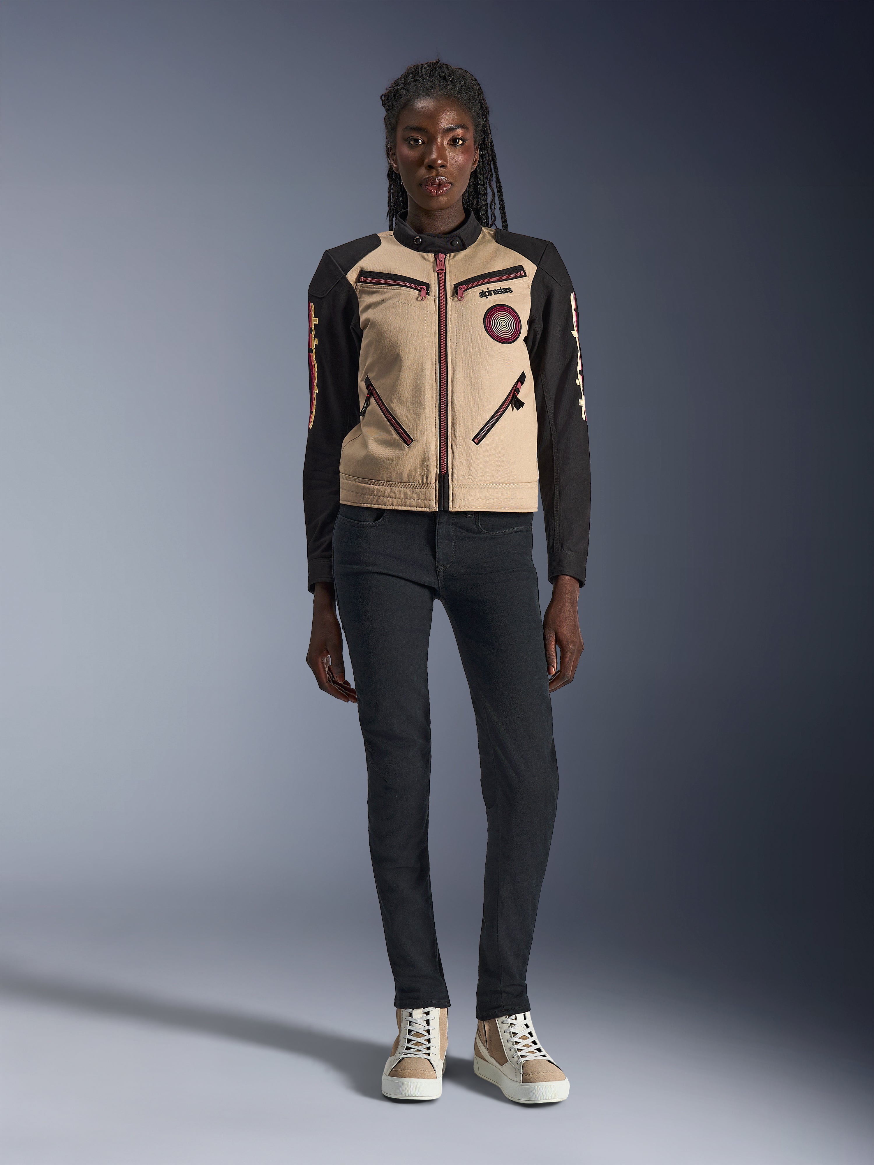 Alpinestars Stella Grace damesmotorjas, stedelijke motorjas, beige en zwarte textielconstructie, voorzien van ritsen aan de voorzijde en geborduurde Oscar-spiraalpatch, gedragen met een zwarte broek en hoge sneakers, eigentijdse op werkkleding geïnspireerde motoruitrusting
