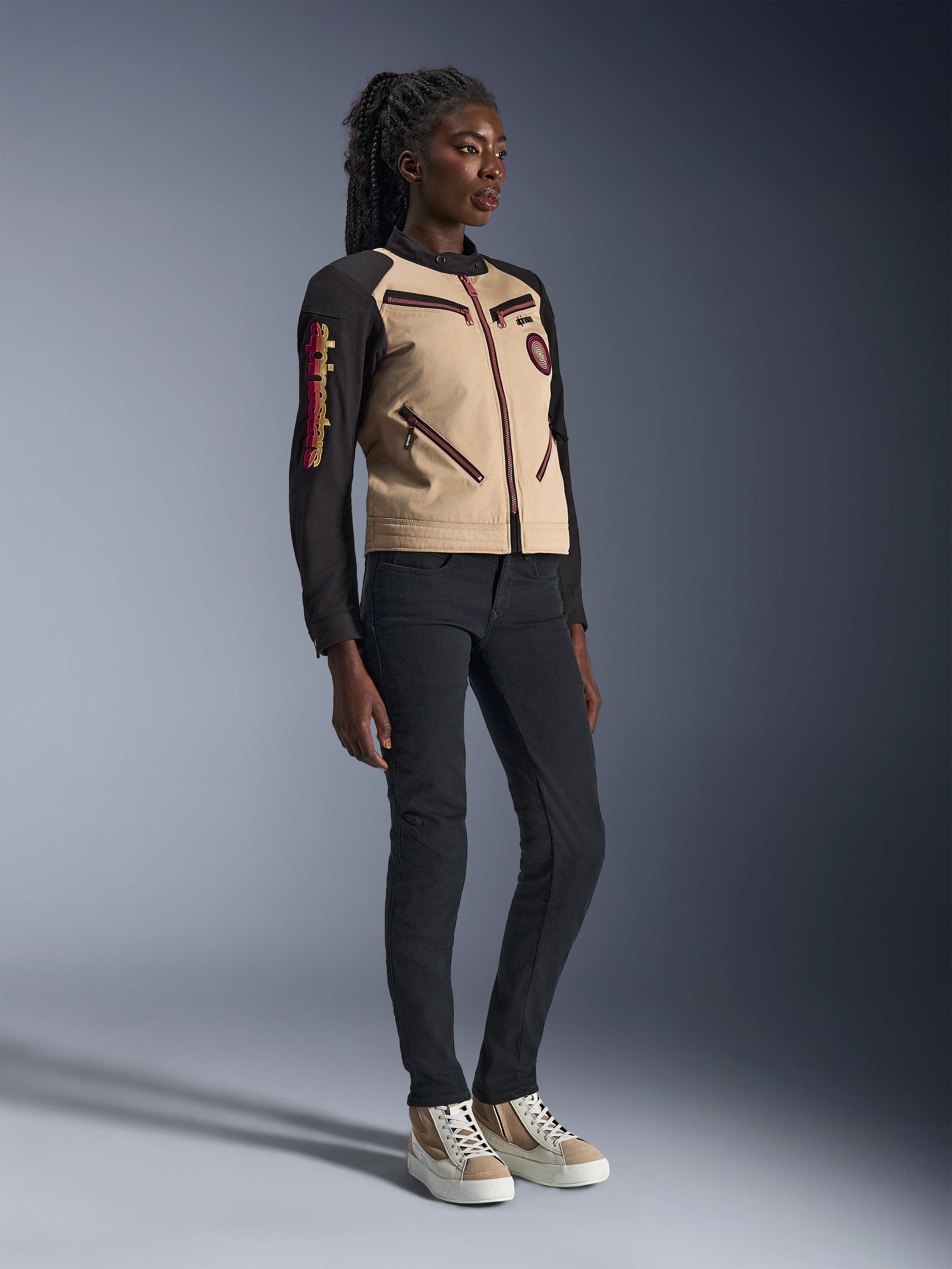Alpinestars Stella Grace jas in Aluminum Black, model draagt jas met beige romp en zwarte mouwen met rode ritsaccenten, gecombineerd met een zwarte slim-fit broek en crèmekleurige sneakers, urban eigentijdse motorkleding voor dames