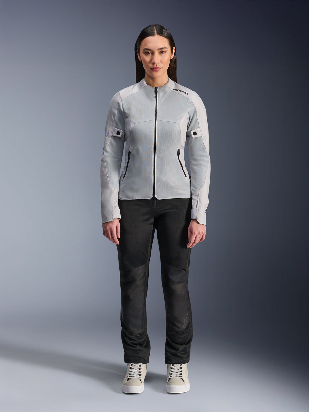 Vrouw die de Alpinestars Stella C-1 Air Jas in zilver draagt, vooraanzicht, urbane motorjas van textiel met tech mesh panelen, gecombineerd met een zwarte broek en witte sneakers, eigentijdse motoruitrusting