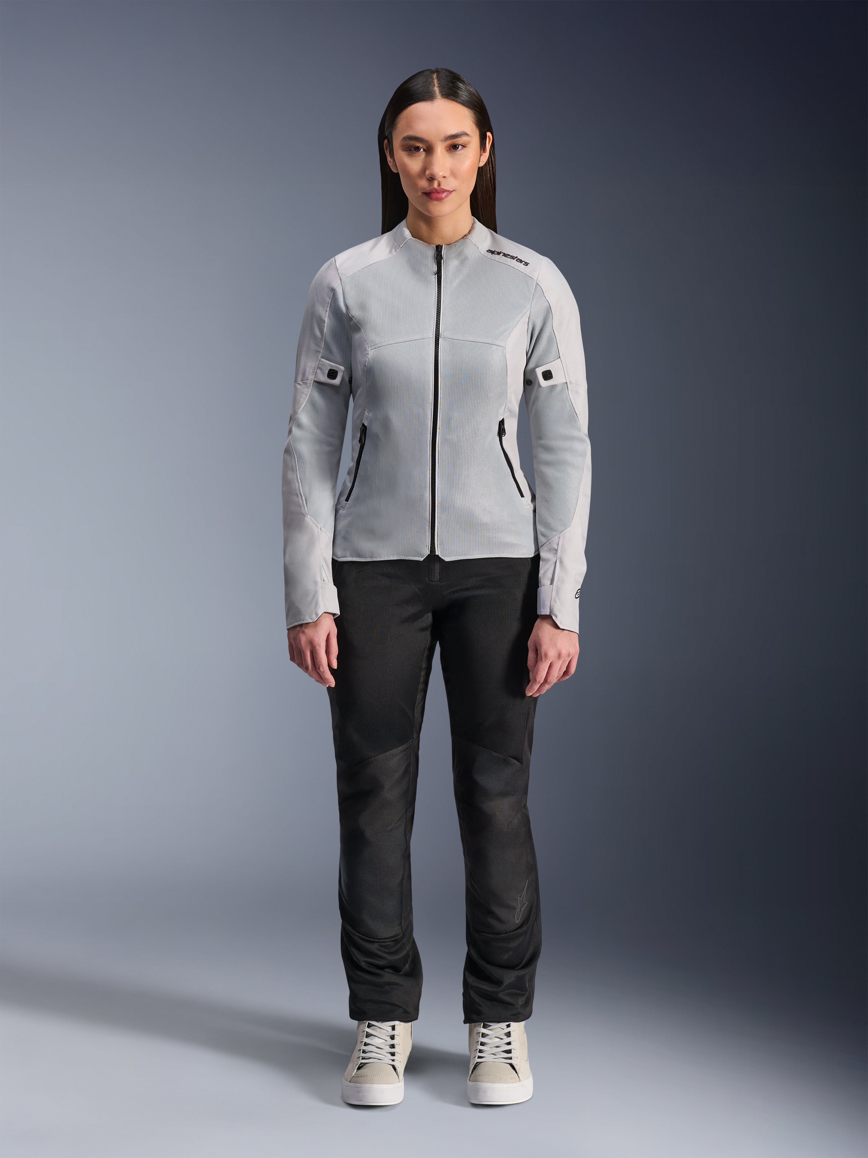 Vrouw die de Alpinestars Stella C-1 Air Jas in zilver draagt, vooraanzicht, urbane motorjas van textiel met tech mesh panelen, gecombineerd met een zwarte broek en witte sneakers, eigentijdse motoruitrusting
