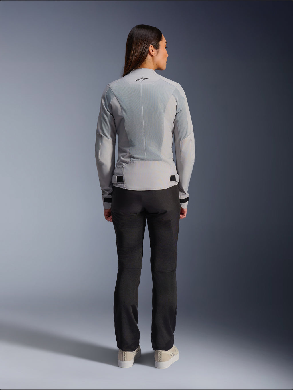 Vrouw draagt Alpinestars Stella C-1 Air Jacket in Silver, achteraanzicht, met grote tech mesh-panelen voor ventilatie en een reflecterend ster-logo, gecombineerd met een zwarte motorbroek en witte sneakers, urban hedendaagse motoruitrusting