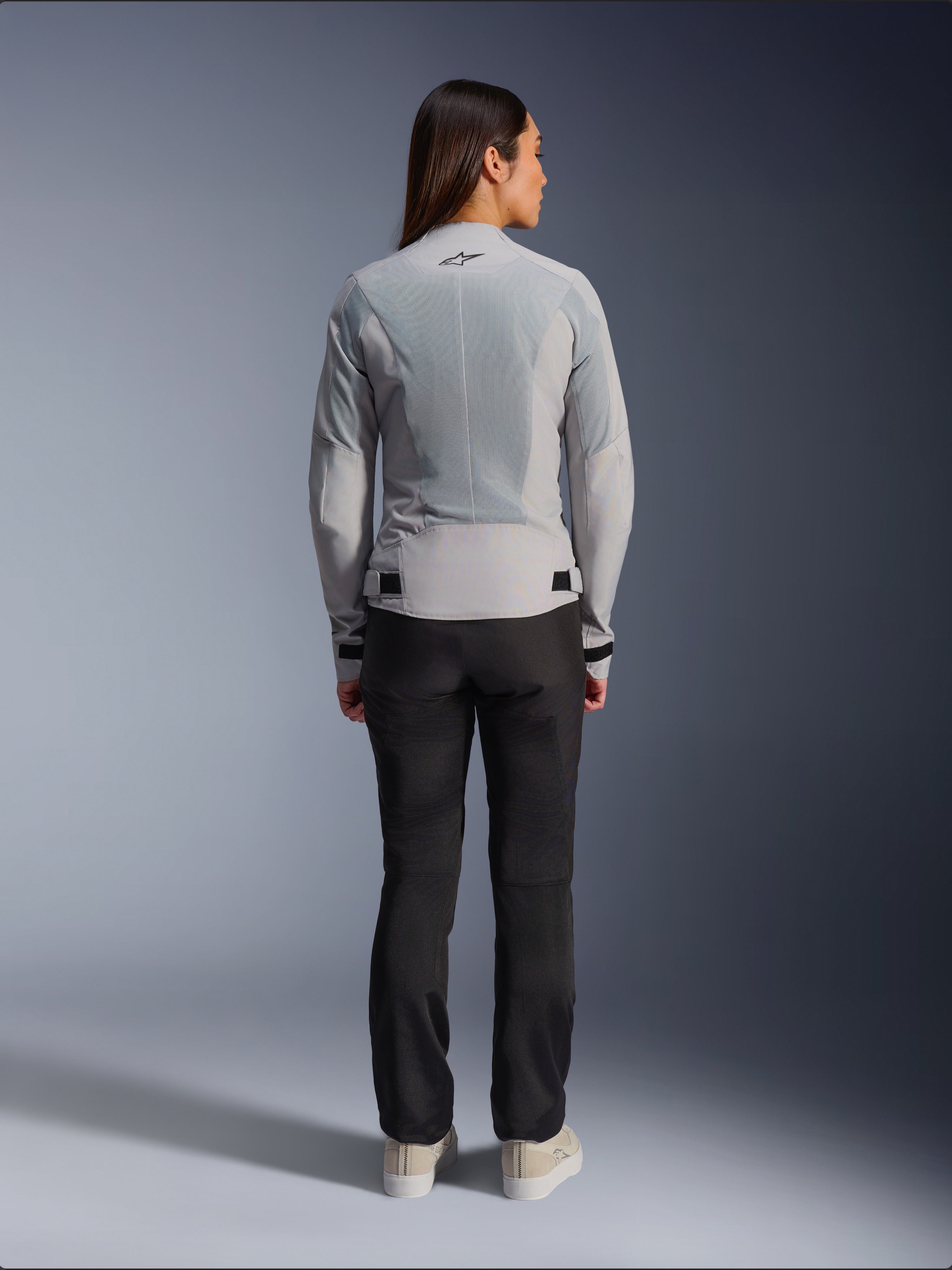Vrouw draagt Alpinestars Stella C-1 Air Jacket in Silver, achteraanzicht, met grote tech mesh-panelen voor ventilatie en een reflecterend ster-logo, gecombineerd met een zwarte motorbroek en witte sneakers, urban hedendaagse motoruitrusting