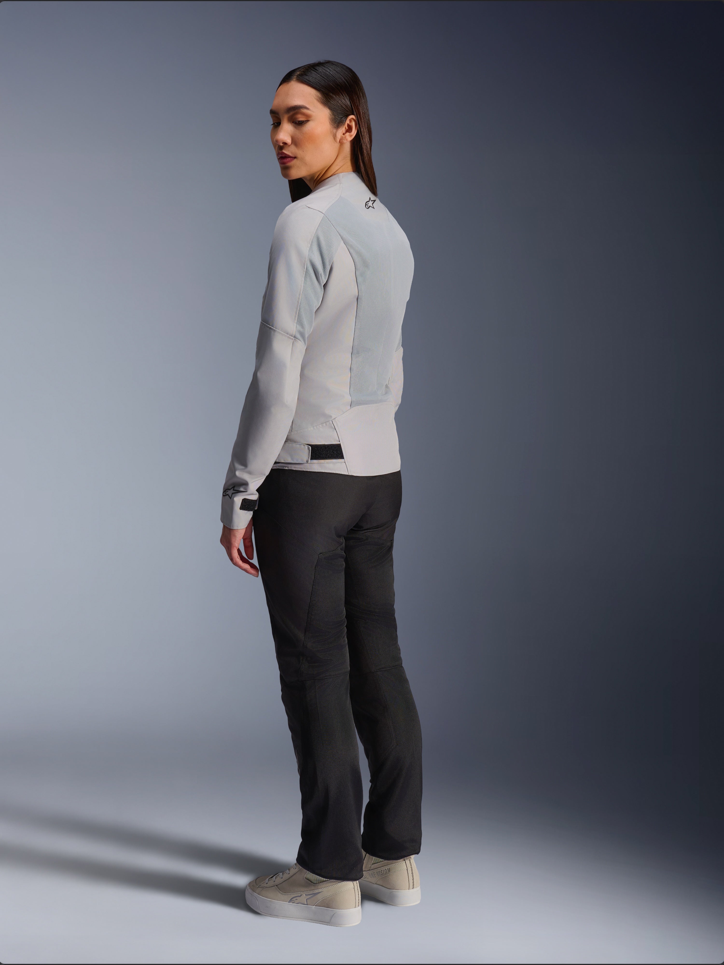 Vrouw die de Alpinestars Stella C-1 Air Jas in Silver draagt, achter- en zijaanzicht met tech mesh-panelen en reflecterend logo op de schouder, gecombineerd met een zwarte motorbroek en beige sneakers, eigentijdse urban rijstijl
