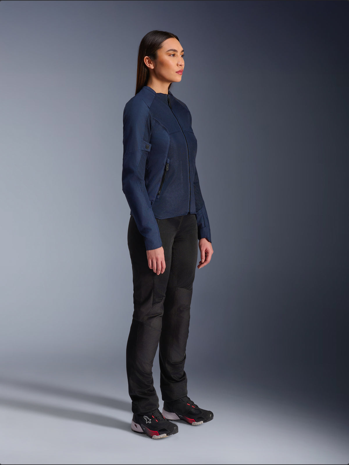 Vrouw in de blauwe Alpinestars Stella C-1 Air Jacket, urban motorjas met lichtgewicht mesh-panelen voor ventilatie, gecombineerd met een zwarte motorbroek en technische schoenen, slank profiel en verfijnde branding, studio zijaanzicht
