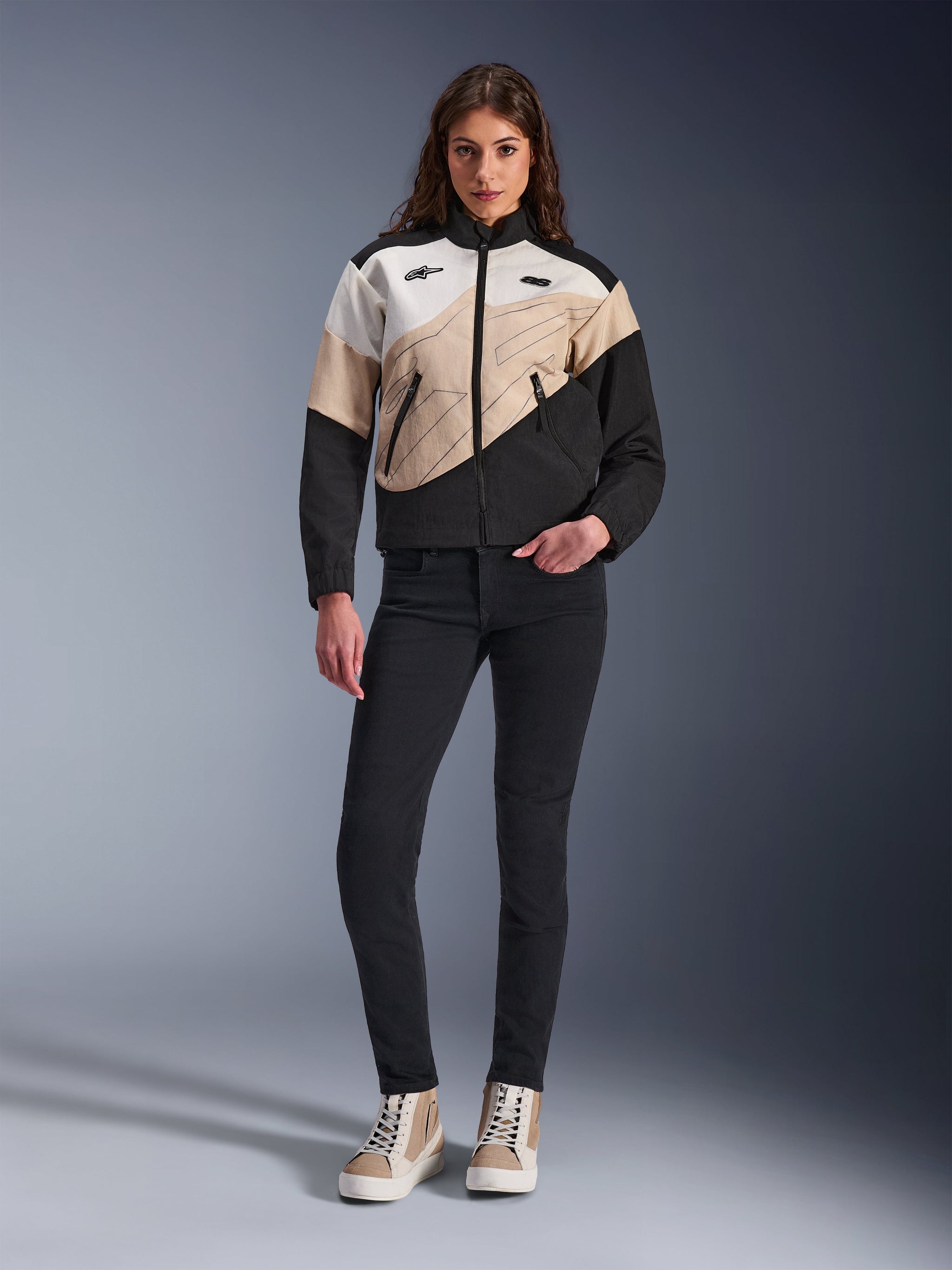 Alpinestars Stella Moshell damesmotorjas, urban motorjas met een asymmetrisch kleurvlakontwerp in zwart, zand en gebroken wit, gedragen met een zwarte slim-fit broek en lichtbruine hoge sneakers, moderne streetwear-esthetiek voor motorrijders.