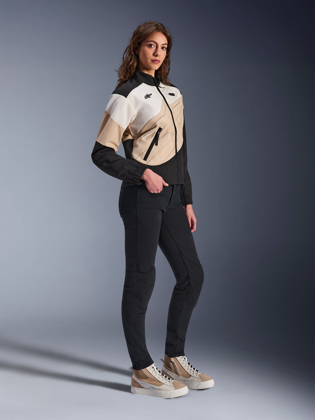 Alpinestars Stella Moshell Jacket, urban motorjas voor dames, zwart, zandkleur en gebroken wit, asymmetrisch geometrisch ontwerp, gedragen met een smalle zwarte broek en beige sneakers, volledige studio-opname van eigentijdse stedelijke motorkleding