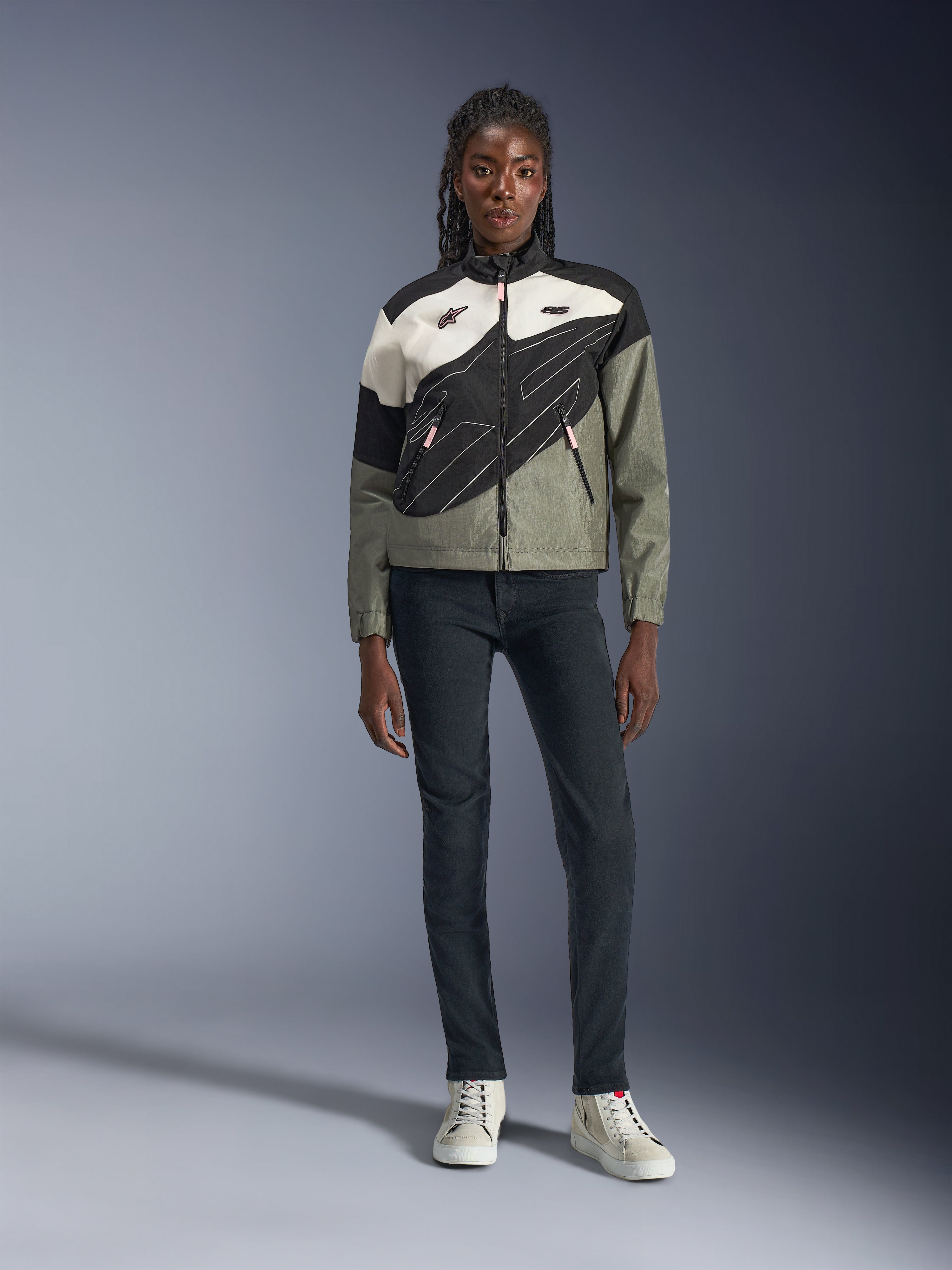 Alpinestars Stella Moshell motorjas, dames motorjas, zwart, grijs en gebroken wit met roze accenten, gedragen door een model met een donkere jeans en gebroken witte sneakers, voorzien van een asymmetrisch ritsontwerp met opvallende geometrische graphics, eigentijdse urban motoruitrusting