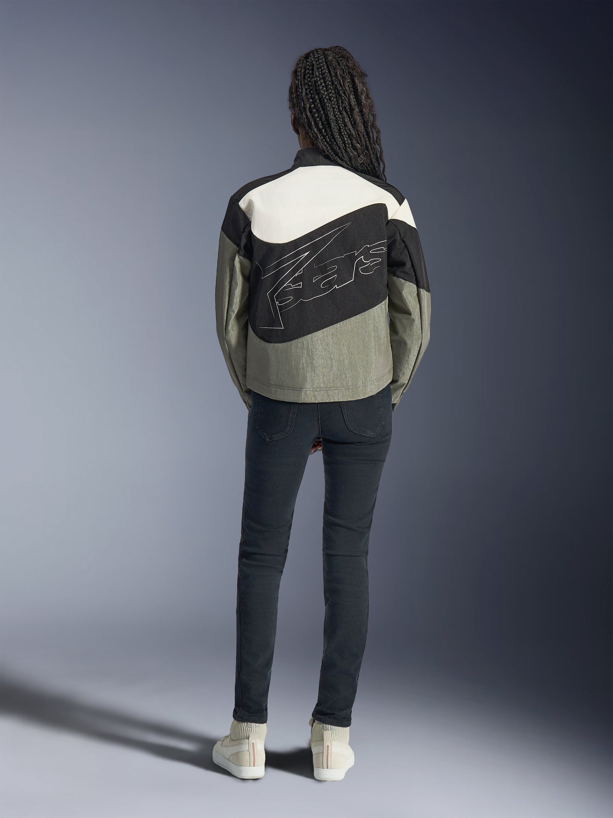 Achteraanzicht van een model in de Alpinestars Stella Moshell Jas in zwart, grijs en gebroken wit, voorzien van een asymmetrisch ontwerp en groot gestileerd logo op de rug, gecombineerd met een donkere slim-fit jeans en witte sneakers, op urban streetwear geïnspireerde motorkleding voor dames