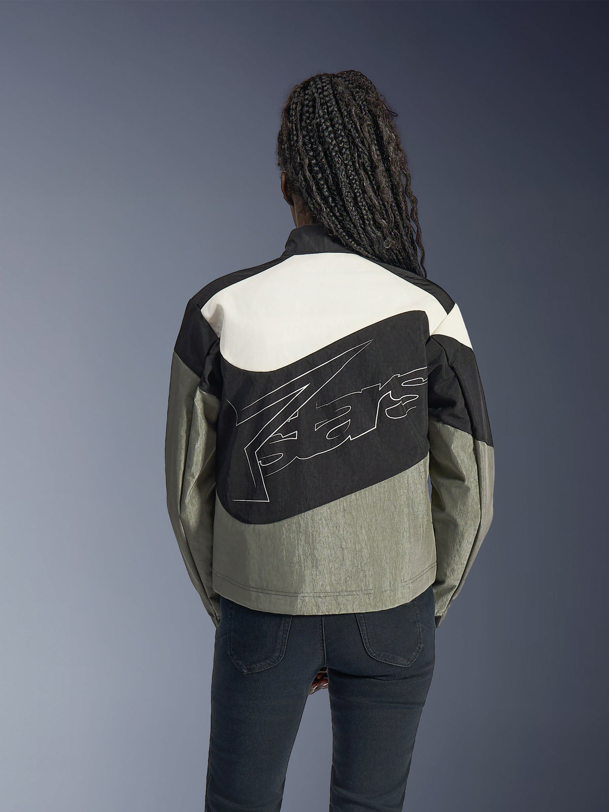 Vrouw van achteren gezien in de Alpinestars Stella Moshell Jas, zwarte, grijze en gebroken witte urban motorjas, voorzien van een groot wit omlijnd logo op de rug, gecombineerd met een zwarte broek, eigentijdse op streetwear geïnspireerde motorkleding.